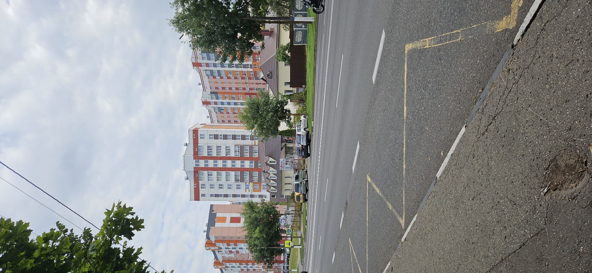 A street scene in Vitsebsk, Belarus, features a multi-lane asphalt road with white and yellow lane markings. In the foreground, the road surface is cracked and weathered, with some debris on the shoulder. Across the road, a row of modern apartment buildings with colorful facades in orange, white, and gray dominates the background, interspersed with trees. In front of the apartment buildings, there is a smaller, older building with a pitched roof, possibly a commercial establishment, identified by signage that reads "ARITEX". Several cars are parked along the side of the road, and a motorcycle is visible further down the street. Trees line the sidewalk in front of the buildings. The sky is overcast with light clouds.