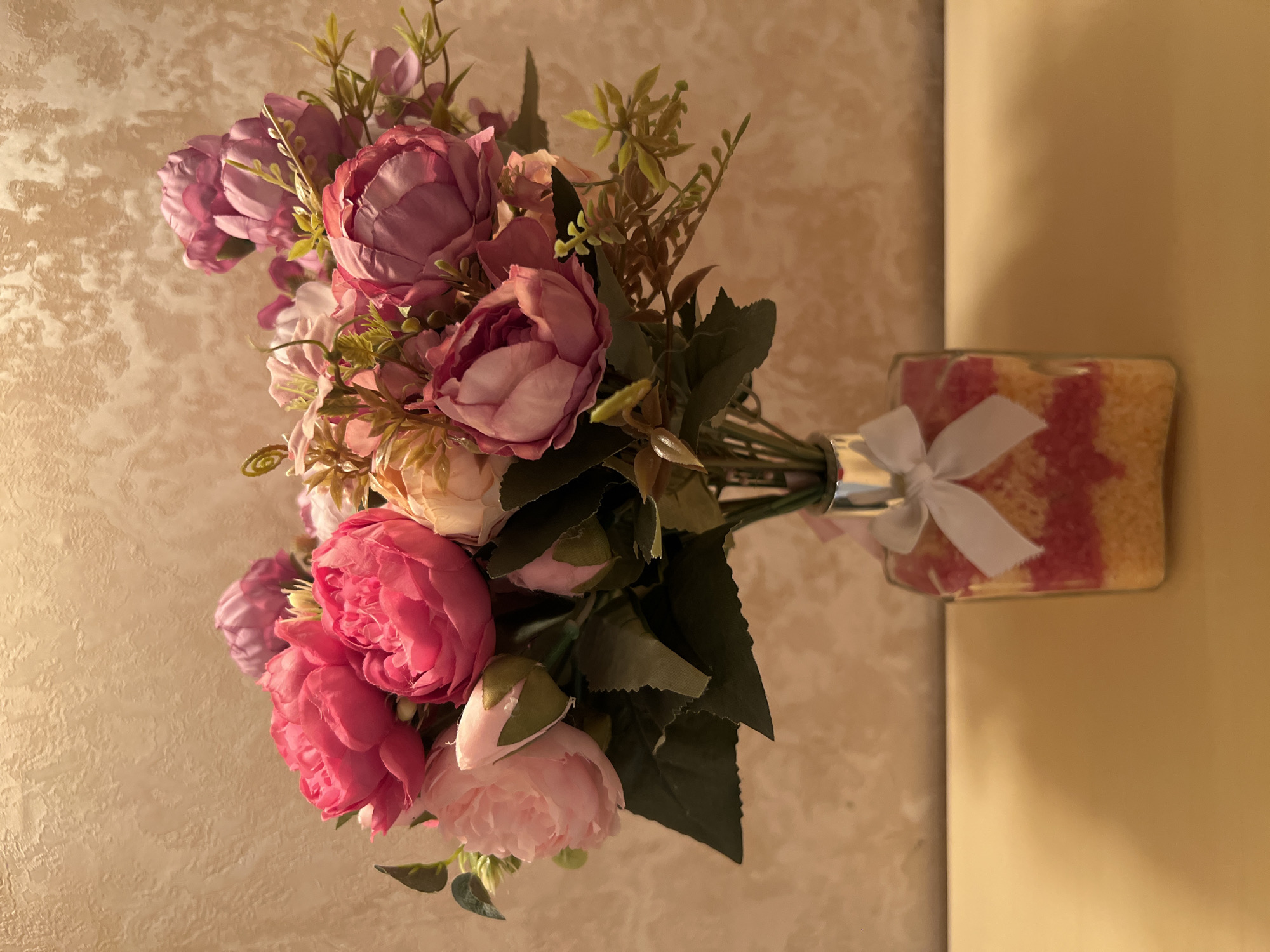 A bouquet of artificial pink and purple peonies is displayed in a clear glass bottle filled with layered pink and yellow decorative beads. The bottle is tied with a white ribbon. The arrangement sits on a light brown wooden surface against a textured, beige wall. There are no people present in the image. The lighting is warm and appears to be coming from above and to the right. The scene is indoors, likely in a residential or decorative setting. No specific location in the city of Bender, Moldova, can be determined from the visual information provided.