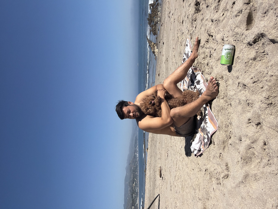 An adult male is seated on a sandy beach in Malibu, United States of America, under a clear blue sky. He is shirtless, wearing dark swim shorts, and positioned on a patterned towel (white, orange, and black). He embraces a medium-sized, brown, curly-haired dog, which has its tongue slightly extended. The man's bare feet are visible in the sand. To his right, a green and white "Spindrift" brand beverage can rests upright on the sand. In the background, the Pacific Ocean shows gentle waves, with dark rock formations visible in the water on the right. Distant, hazy coastal hills line the horizon to the left, showing signs of development. A portion of a black tripod or stand is visible on the far left edge of the foreground.