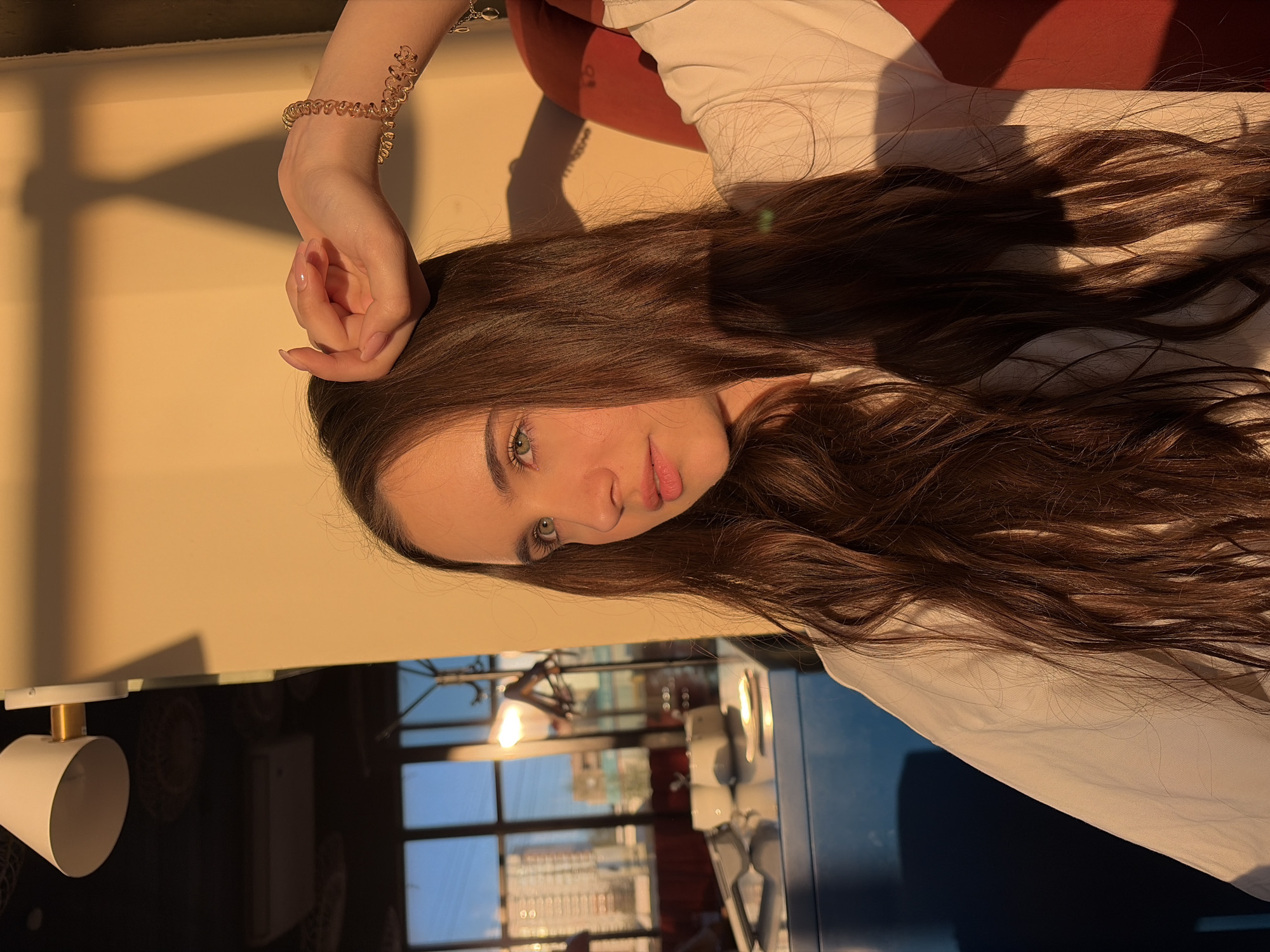 A young woman with long brown hair and green eyes is posing indoors. Her left hand is raised near her head, showcasing a gold bracelet. She wears a white shirt and is partially reclined on an orange surface. The backdrop features a window view of Izhevsk, Russia. Visible through the window are building structures and a blue sky, hinting at an urban setting. In the foreground, parts of a kitchen or dining area with blue-colored furniture are visible, as well as a lamp mounted to a ceiling.