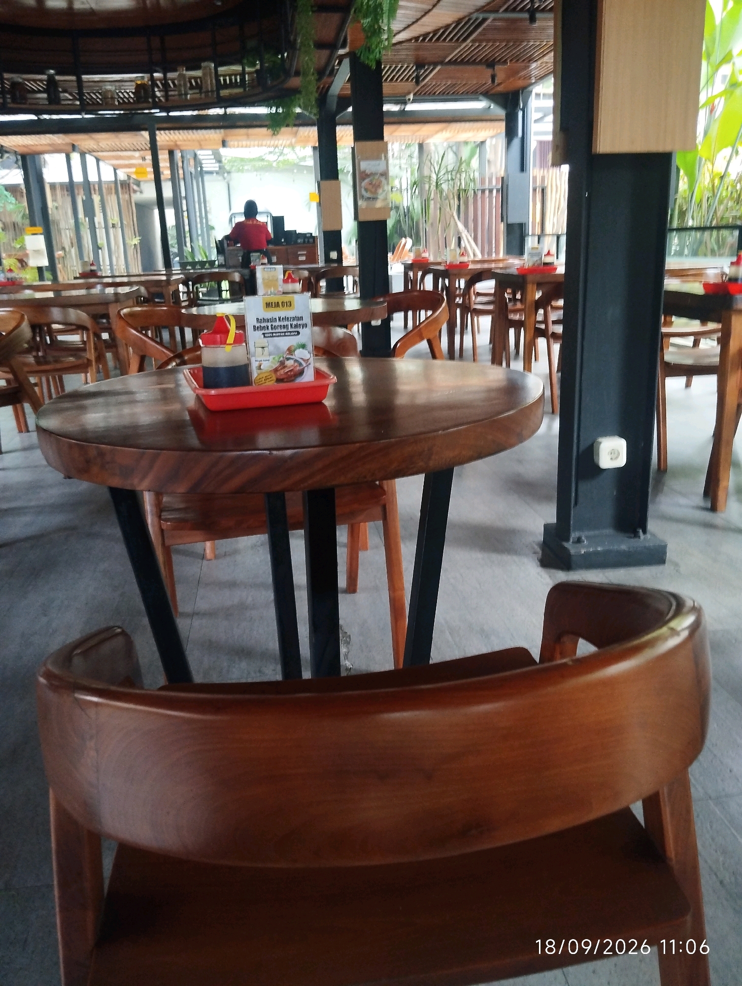 The image shows the interior of a restaurant with a rustic, open-air design, featuring wooden furniture and ceiling accents. In the foreground, a round wooden table with black metal legs is visible, set with a red tray containing a bottle of dark sauce and a smaller bottle with a yellow cap. A close-up of a wooden chair back is in the bottom foreground. In the background, other tables and chairs are arranged, and a person wearing a red shirt is standing near a counter. The restaurant appears to be in a warm climate, with greenery visible through large windows and along the ceiling. The timestamp at the bottom right indicates "18/09/2026 11:06". The location is stated to be in the city of Bandung City, Indonesia.