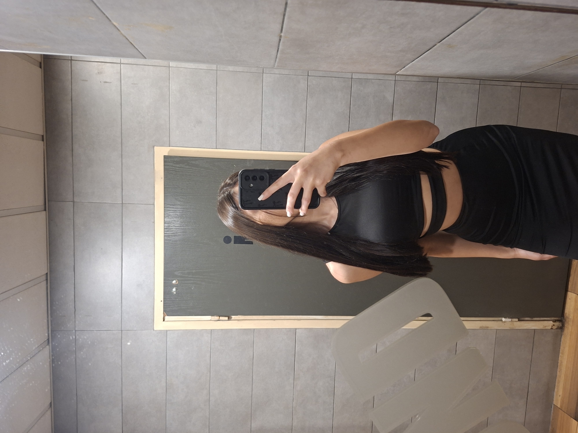 A woman in a black two-piece dress is taking a mirror selfie in a public restroom with gray tiled walls. The stall door is visible, and a reflective surface with obscured markings is present. The location appears to be a restroom, possibly in Pleven, Bulgaria, though this is not explicitly confirmed.