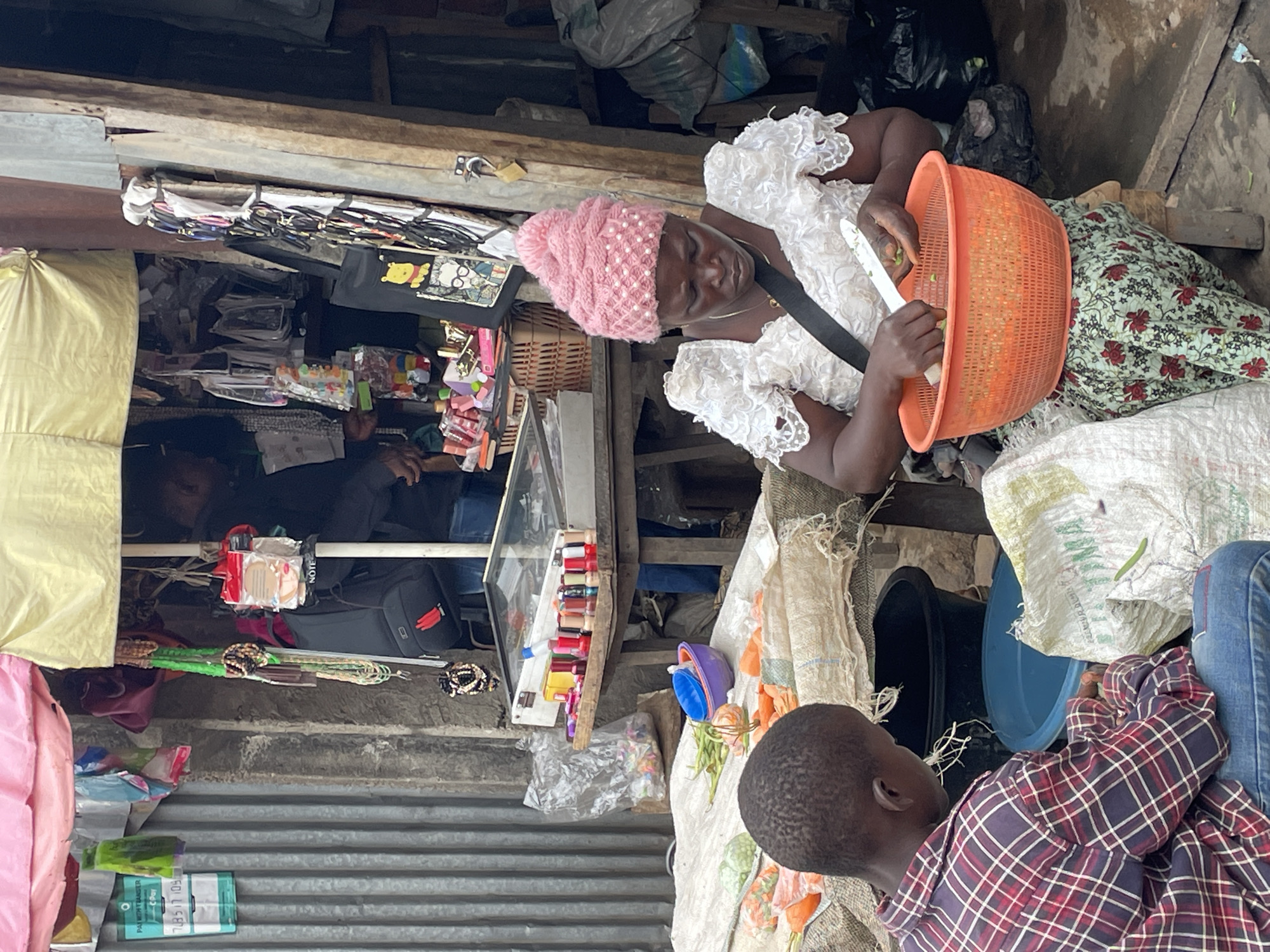 An outdoor scene in Jos, Nigeria, features two individuals and various objects associated with commerce and daily life. An adult woman with dark skin and a pink, pearl-adorned knitted hat is shown wearing a white lace top and a black strap across her chest. She sits with an orange plastic basket on her lap, and she is cutting green vegetables inside the basket with a white utensil. A child is shown, wearing a red and blue plaid shirt and blue jeans, looking toward a blue bucket.
Behind the woman, there appears to be a makeshift stall or shop with a variety of items displayed, including jewelry, bags, and cosmetic items. The stall has a corrugated metal backing. Bags of items are stored above the stall. The environment seems cramped and possibly part of a market area or street-side setup.
