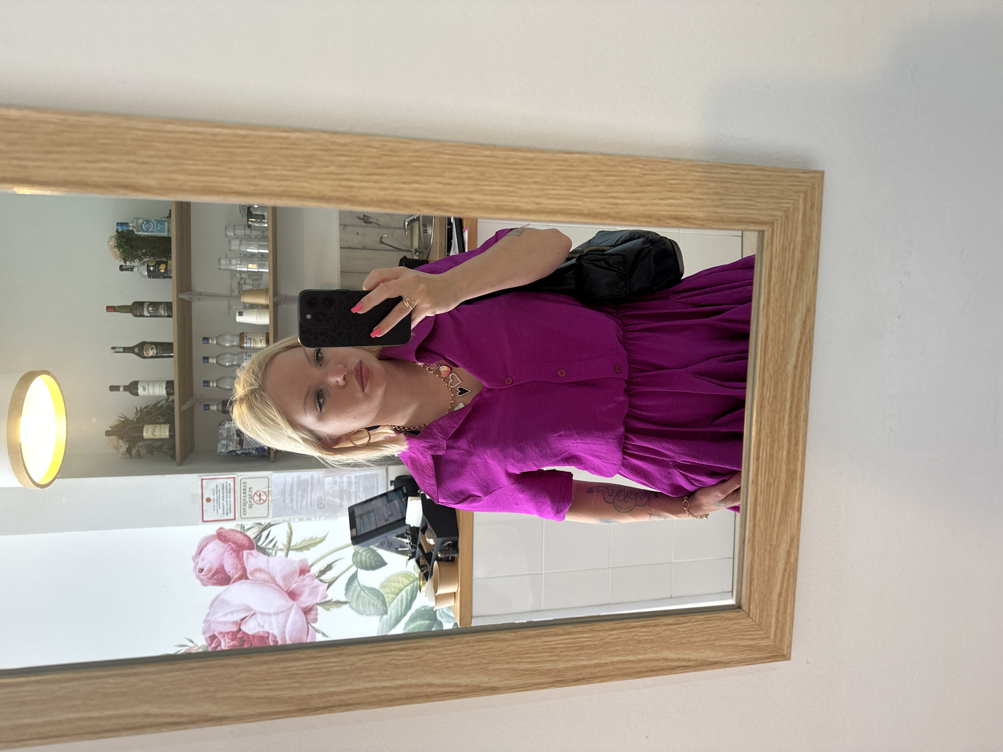 The image depicts a mirror selfie taken indoors within an establishment in Budva, Montenegro. A light-skinned blonde woman, appearing to be in her 20s or 30s, is reflected in a large mirror with a natural wood frame. She has winged eyeliner, light pink lipstick, and wears large gold hoop earrings. Her short blonde hair is styled. She is wearing a vibrant magenta-colored collared dress with short sleeves and a cinched waist. A prominent gold chain necklace with multiple colorful heart-shaped pendants (gold, pink, green, white) adorns her neck. A gold-toned bracelet is visible on her right wrist, and a coiled tattoo is present on her left forearm. Her fingernails are painted bright pink. She holds a dark-colored smartphone, likely an iPhone, in her right hand, positioned to capture the mirror reflection. A black quilted shoulder bag is partially visible on her left side.
The background, also reflected in the mirror, shows the interior of what appears to be a cafe or bar. To the left of the woman, a white wall features a large mural of pink roses with green leaves. Below the mural, a counter area with two light brown disposable cups and a black point-of-sale (POS) system is visible. To the right of the woman, a white wall holds two wooden shelves. The top shelf displays several bottles of alcohol, including wine and spirits, and a small floral arrangement. The middle shelf holds various drinking glasses and small cups. Below the shelves, a white tiled backsplash is visible, leading to a counter with a sink and faucet on the far right. A circular light fixture with a warm glow is visible on the ceiling. Additionally, a white sign with red text and an inverted "no smoking" sign are affixed to the wall near the counter area.