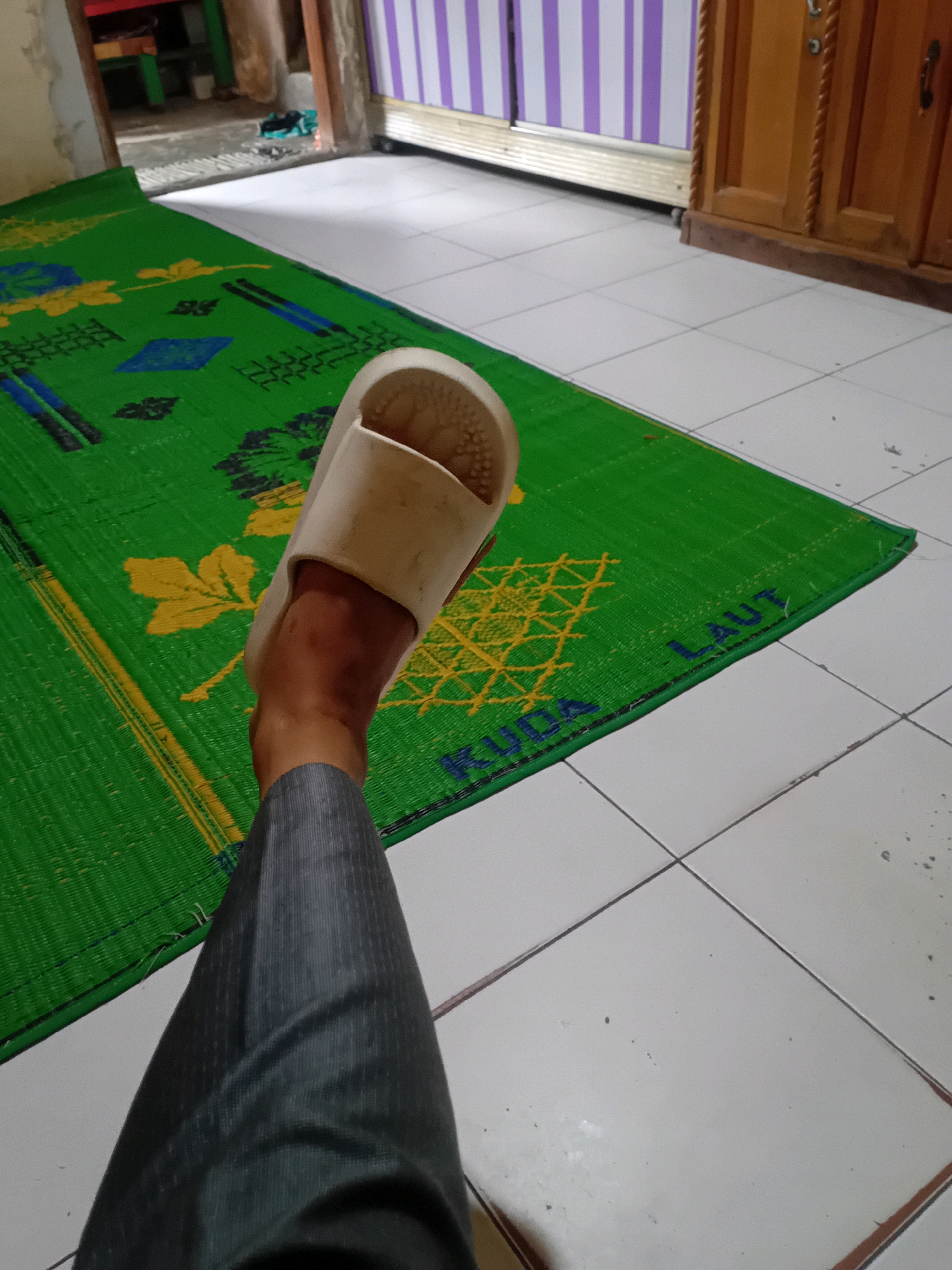 A person's left leg, clad in gray striped trousers, is shown from the thigh down, with a white slipper on their foot. The leg is positioned on a green woven mat with a yellow floral pattern and blue diamond shapes. The mat is laid on a white tiled floor. In the background, a wooden cabinet with a textured finish is visible. To the left of the cabinet, a doorway leads to another room with striped purple and white curtains. The scene appears to be indoors, likely in a residential setting in Pekanbaru, Indonesia.
