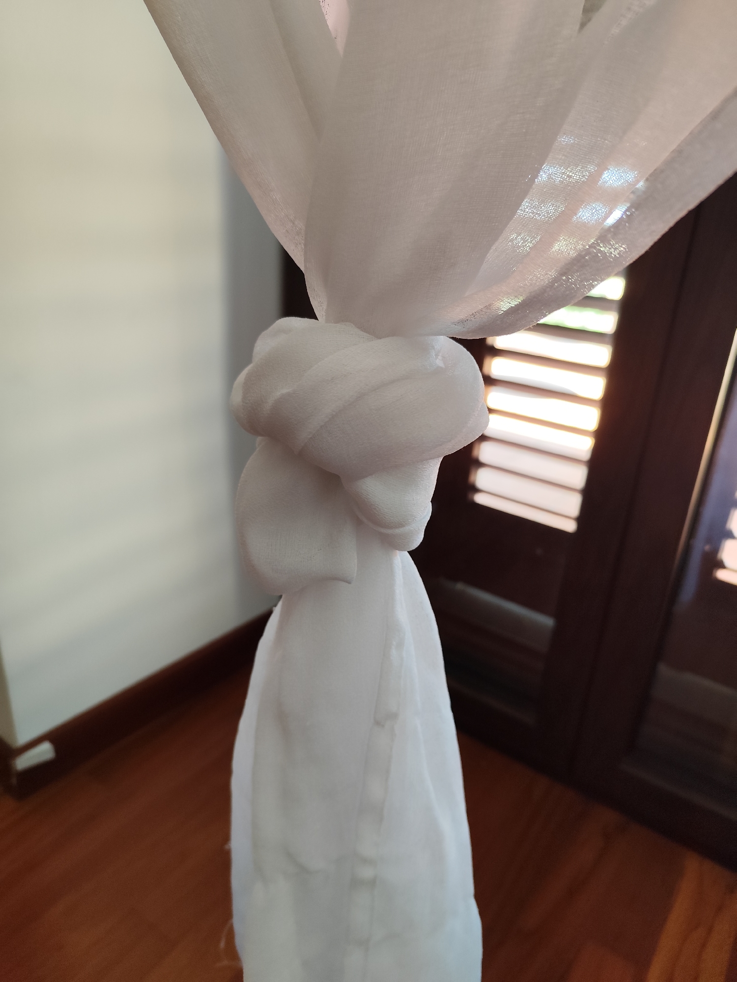 A close-up shows a knot tied in a white, semi-sheer curtain. The curtain is tied against a backdrop of dark brown wooden shutters. The light coming through the shutters creates horizontal stripes. The wooden floor is visible in the foreground. A pale wall is partially visible on the left side of the frame. The location is not identifiable as Giarre, Italy from the visual content of the image.