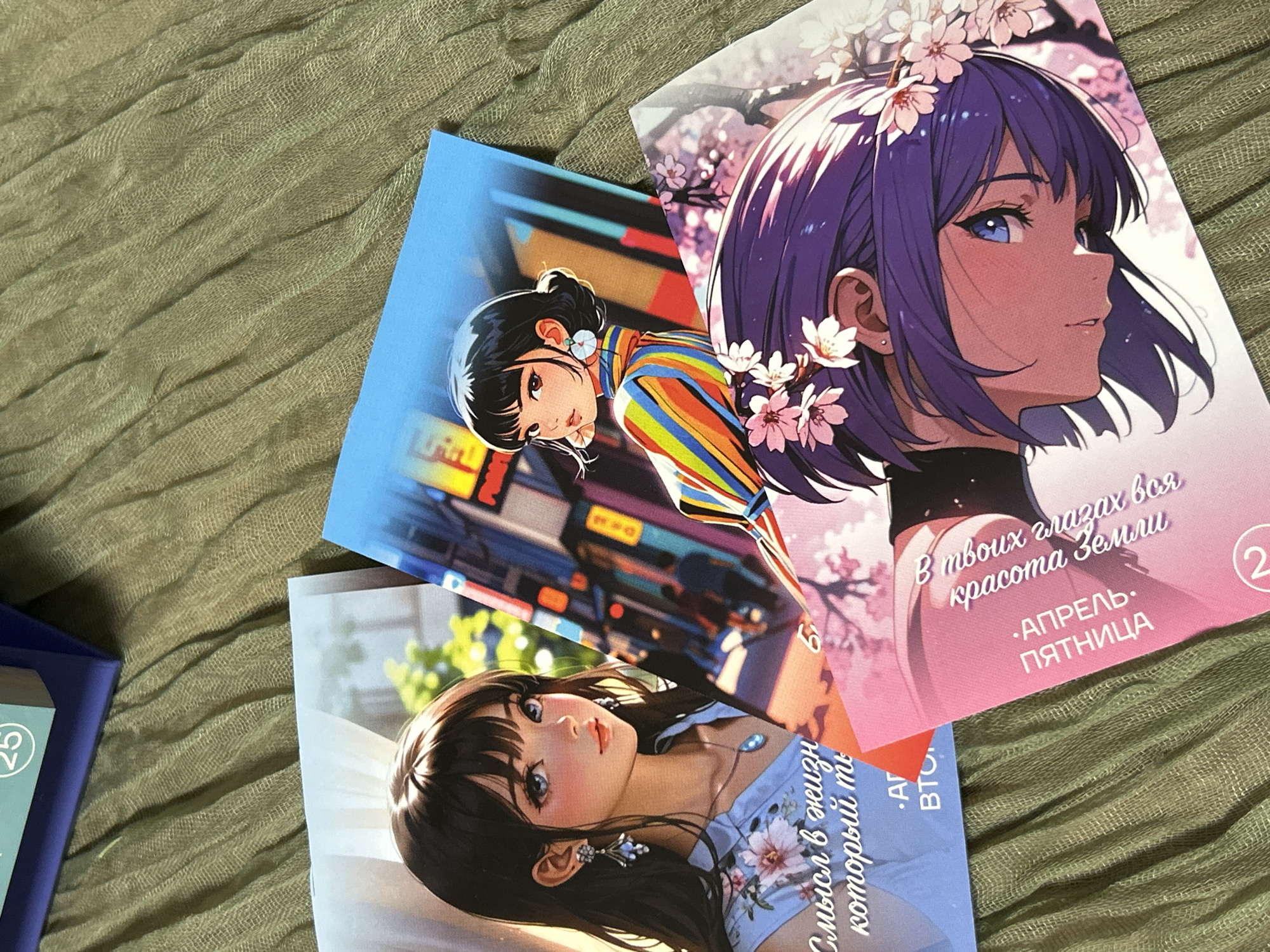 The image shows three postcards with anime-style illustrations of young women. The postcards are arranged haphazardly on a textured, olive-green surface, likely a piece of fabric or a tablecloth.
The postcard on the top right features a girl with short, purple hair adorned with cherry blossoms, depicted in profile. Text in Russian is visible on the lower part of the postcard.
The postcard in the middle depicts a girl with black hair, wearing a vibrant, striped top. The background suggests a city setting, with blurred lights and buildings.
The postcard on the bottom left showcases a girl with long, dark hair wearing a light blue dress with floral patterns. She also has earrings. Some Russian text is visible on the postcard.
A glimpse of a book or another item with the number "25" on its spine is visible on the left side of the image.
Based on the Russian text and the style of illustration, it is possible the postcards are related to or were purchased in Russia, but there is no indication of the specific location in Saint Petersburg.