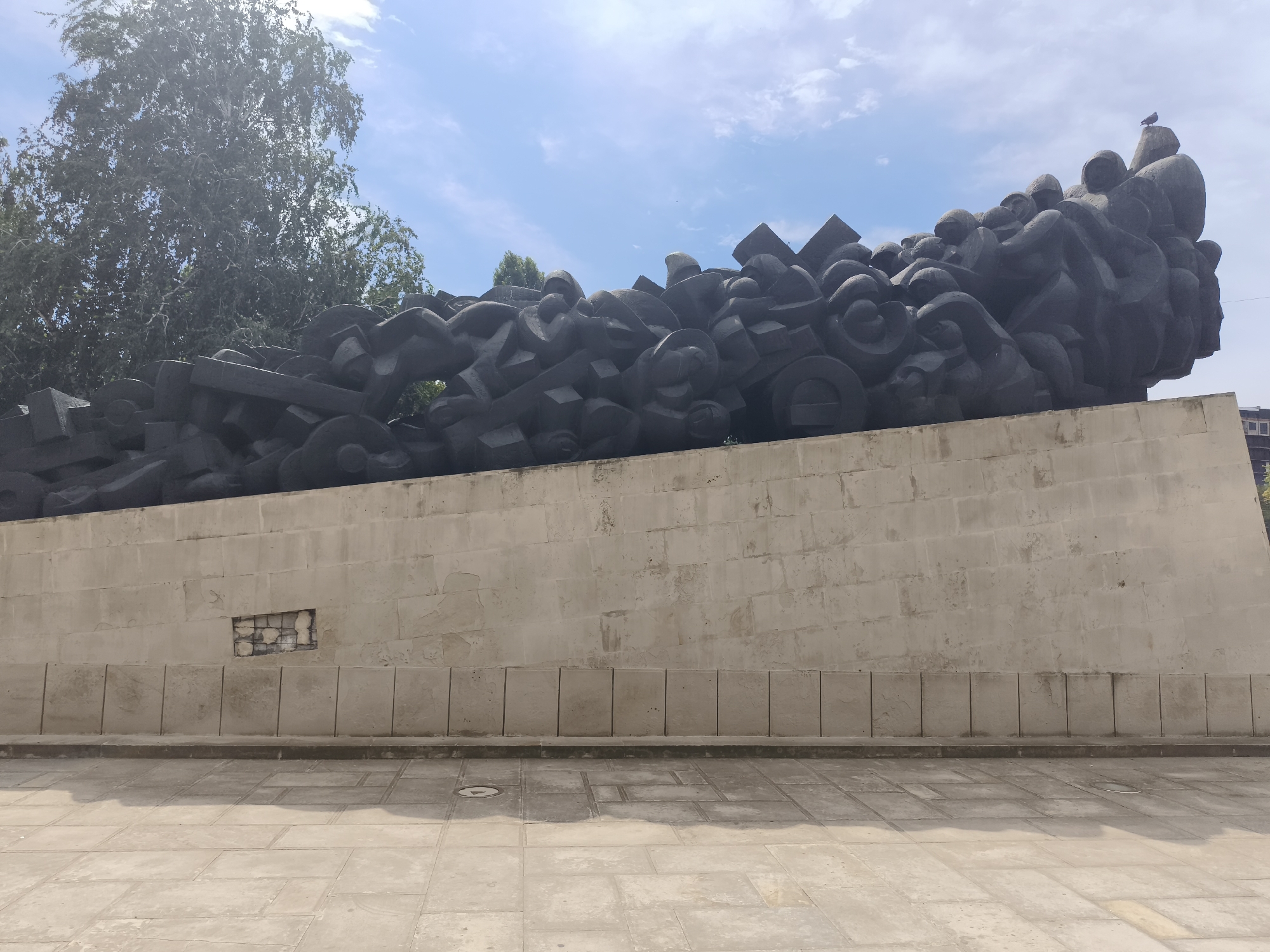 A wide-angle, eye-level shot captures a monumental abstract sculpture in Chișinău, Moldova, under a partly cloudy blue sky. The sculpture, made of dark, rough-hewn stone, is piled high on a massive, light-colored concrete retaining wall. The wall is constructed of large, square blocks and topped with a row of vertical, rectangular stone slabs. The base of the wall meets a paved courtyard, with large, square tiles forming a pattern. A section of the concrete wall near the left side is damaged, revealing a rougher, brick-like interior. Trees with green foliage are visible behind and to the left of the sculpture. A single bird perches atop the highest point of the sculpture. The scene is static and devoid of human activity.
