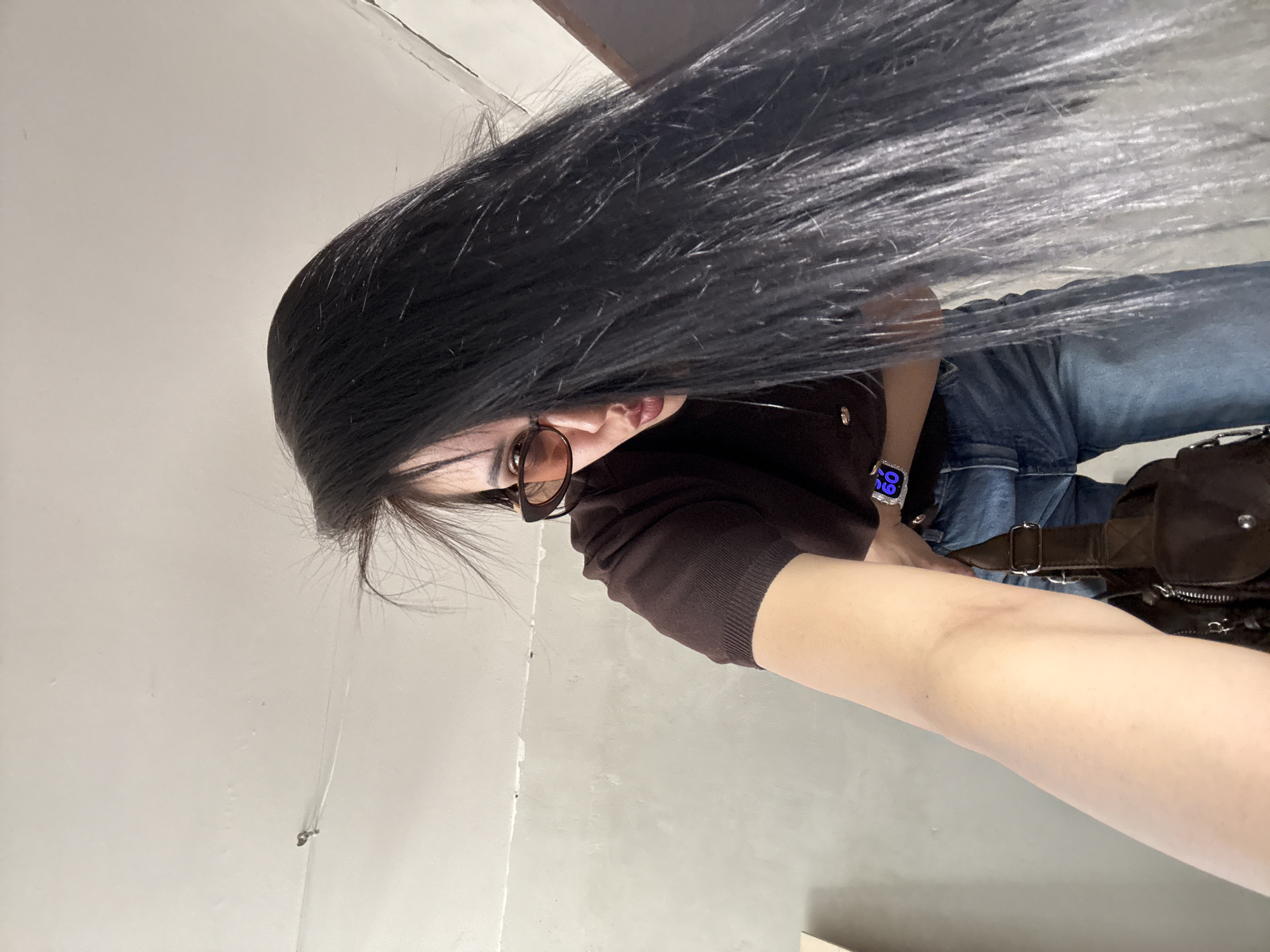 A person with long, straight black hair is taking a selfie indoors. The person is wearing sunglasses, a brown sweater, and blue jeans. A brown bag is slung across their body. The location is not identifiable as being in Votkinsk, Russia. The environment is an interior with plain walls.