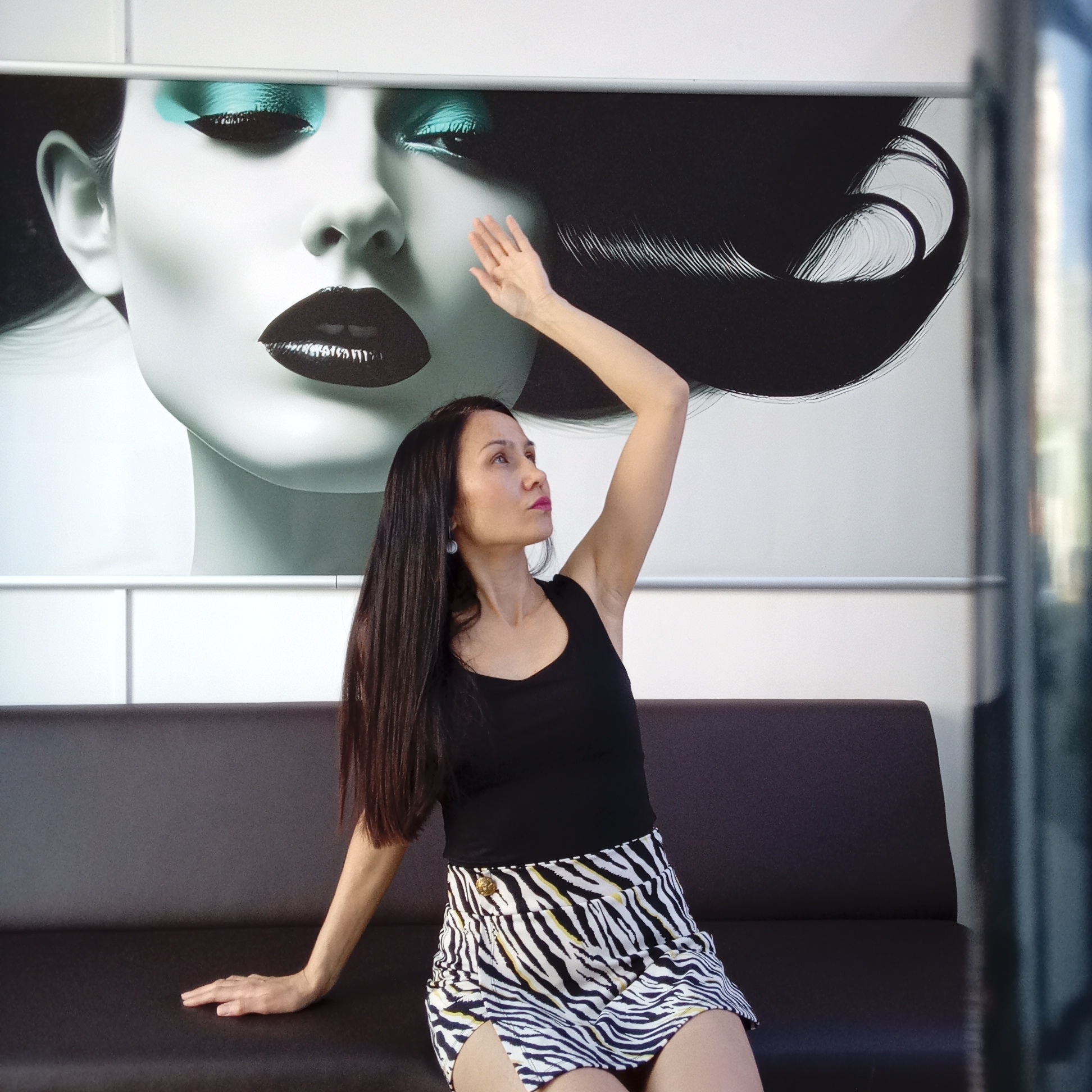 A woman with long dark hair is seated on a dark brown sofa, positioned in front of a large poster of a woman's face. The woman is wearing a black tank top and a zebra-print skirt. Her right arm is raised, hand facing upwards, while her left hand rests on the sofa. The poster depicts a woman with dramatic makeup, including blue eyeshadow and dark lipstick, with stylized dark hair. The location is inferred to be in Izhevsk, Russia.