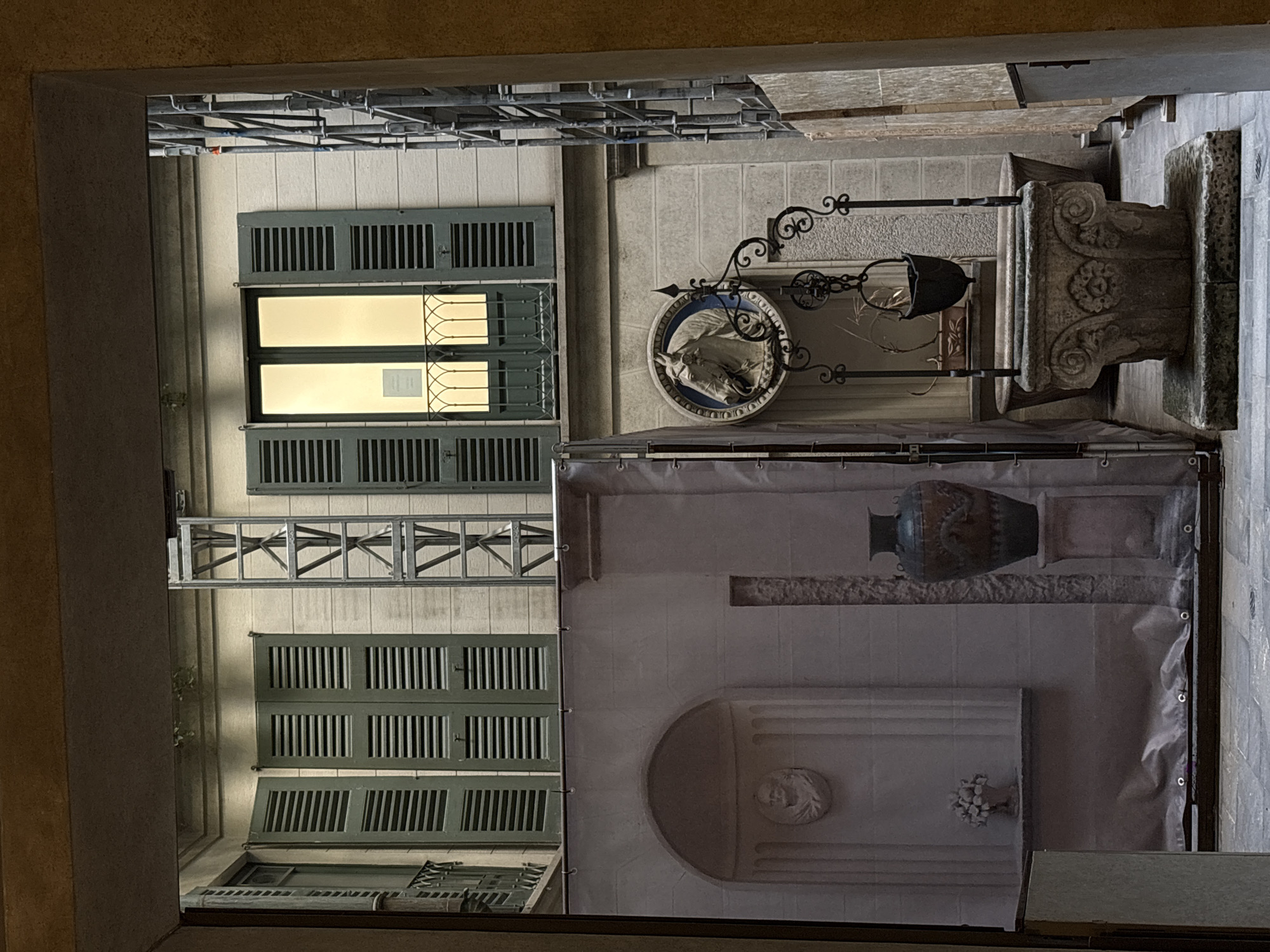 The image, captured from a lower perspective looking upwards, is rotated 90 degrees clockwise. It depicts a view into a dimly lit urban space, possibly a courtyard or narrow passage.
On the left side of the image, running vertically, is the rough-textured inner edge of an archway or opening. Beyond this, a building facade dominates the scene. Towards the upper half of the image, a large window is visible, brightly illuminated from within with a warm yellow light. This window has dark green louvered shutters, both above and below the glass panes. Metal bars with decorative scrollwork cover the window glass, and a small, unreadable white rectangular paper is affixed to one of the panes. Above this window, a complex scaffolding structure is mounted to the building. Another set of dark green louvered shutters is visible further down the building face towards the bottom of the image, along with a glimpse of another window.
On the right side of the image, various decorative and architectural elements are present. Towards the top, an ornate wrought-iron bracket supports a circular sign featuring a white relief of a horse's head against a blue background. Below this, another wrought-iron fixture holds a dark, bell-shaped object, possibly a lantern. Further down and to the right, a heavily carved stone column base with intricate floral and swirling designs stands on a paved surface. A large fabric banner or tapestry covers a significant portion of the right side, displaying a printed image of a classical vase, a bust within a semi-circular niche, and a small floral sculpture, mimicking architectural details.
The overall lighting is low, suggesting either evening or a heavily shaded interior space. No people are visible, and the scene is static. The architectural style, including the shutters and stone work, is consistent with a historic European city like Milan, Italy. The only visible text is unreadable on the paper within the lit window.