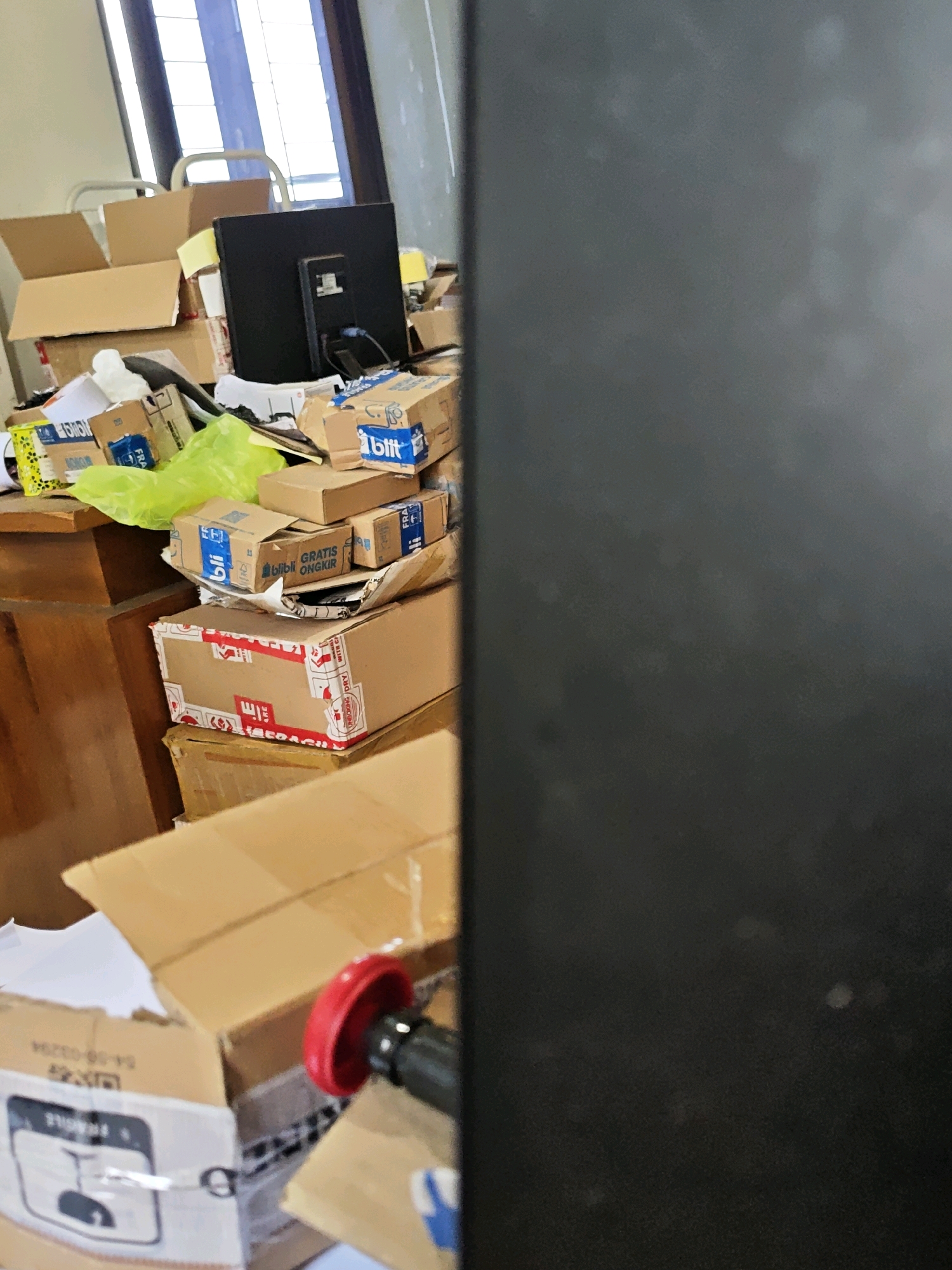In a room in Ngasem, Indonesia, a cluttered desk is piled high with cardboard boxes of various sizes. A black computer monitor sits atop some of the boxes. Many of the boxes have blue and white labels, some of which are partially obscured by other items. A bright green plastic bag is also visible among the boxes. To the right of the desk, a dark, out-of-focus object dominates the frame, possibly a wall or furniture. The overall impression is one of disarray and a significant volume of packages.