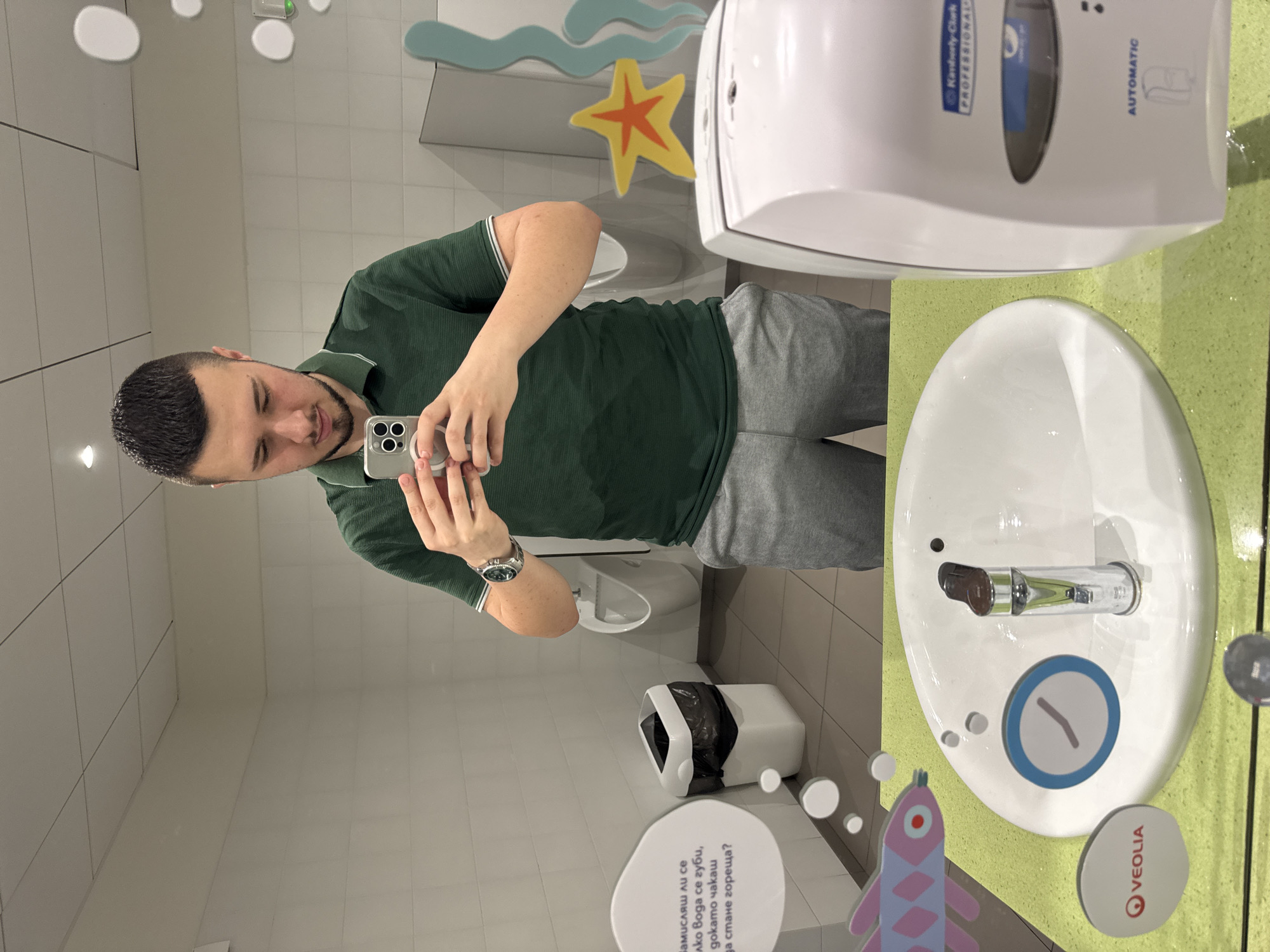 Here is a forensic description of the image:
**Scene:** The image shows a man taking a mirror selfie in what appears to be a public restroom.
**Environment:** The restroom has white tiled walls. There is a child-friendly design to the sink area with colorful fish and sea-themed decals around the sink. The sink is made of white ceramic and has a sensor-operated water faucet. Above the sink is a Kimberly-Clark Professional automatic paper towel dispenser. A trash receptacle is visible, and informational signage with the Veolia logo is present, indicating it could be part of a water-saving campaign.
**Person:** The man is wearing a green polo shirt and grey pants. He is holding a smartphone, presumably to take the selfie.
**Objects:**
* Smartphone with a camera
* Automatic paper towel dispenser
* Ceramic sink with faucet
* Trash receptacle
* Signage with text and logo
* Water-themed decals
**Actions:** The man is taking a selfie.
**Inferred Location:** Based on the text on the informational sign, this location is in Sofia, Bulgaria.
