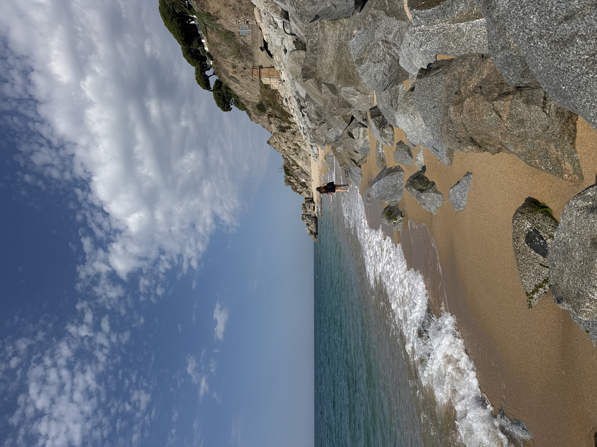 The image depicts a coastal scene in Arenys de Mar, Spain. A young woman with dark hair, wearing a black top and shorts, walks along the shoreline of a sandy beach. She is wading in the shallow, turquoise ocean water. The beach is bordered by large, gray rocks on the right side of the frame, and a rocky, tree-covered cliff rises on the left. The sky above is blue with scattered white clouds. A pathway and some brick structures are visible on the cliff face. The waves are gently rolling onto the shore, creating white foam. The overall impression is of a tranquil summer day at the coast.