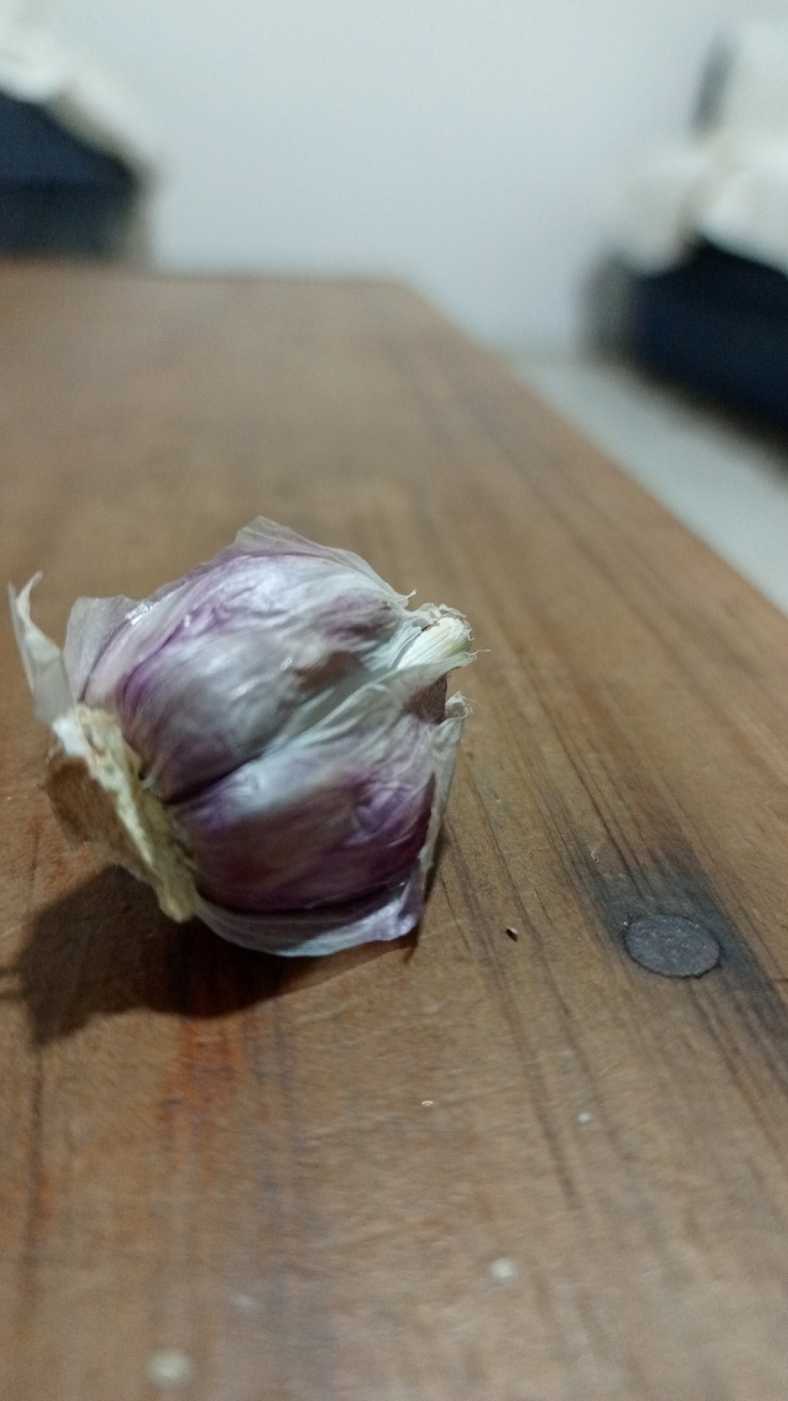 A head of garlic, with purple-tinged cloves visible through its papery skin, rests on a weathered wooden surface. A nail is visible in the wooden surface, and two other objects are visible in the background. There is no indication of this being in Los Robles, Venezuela. No people are visible.