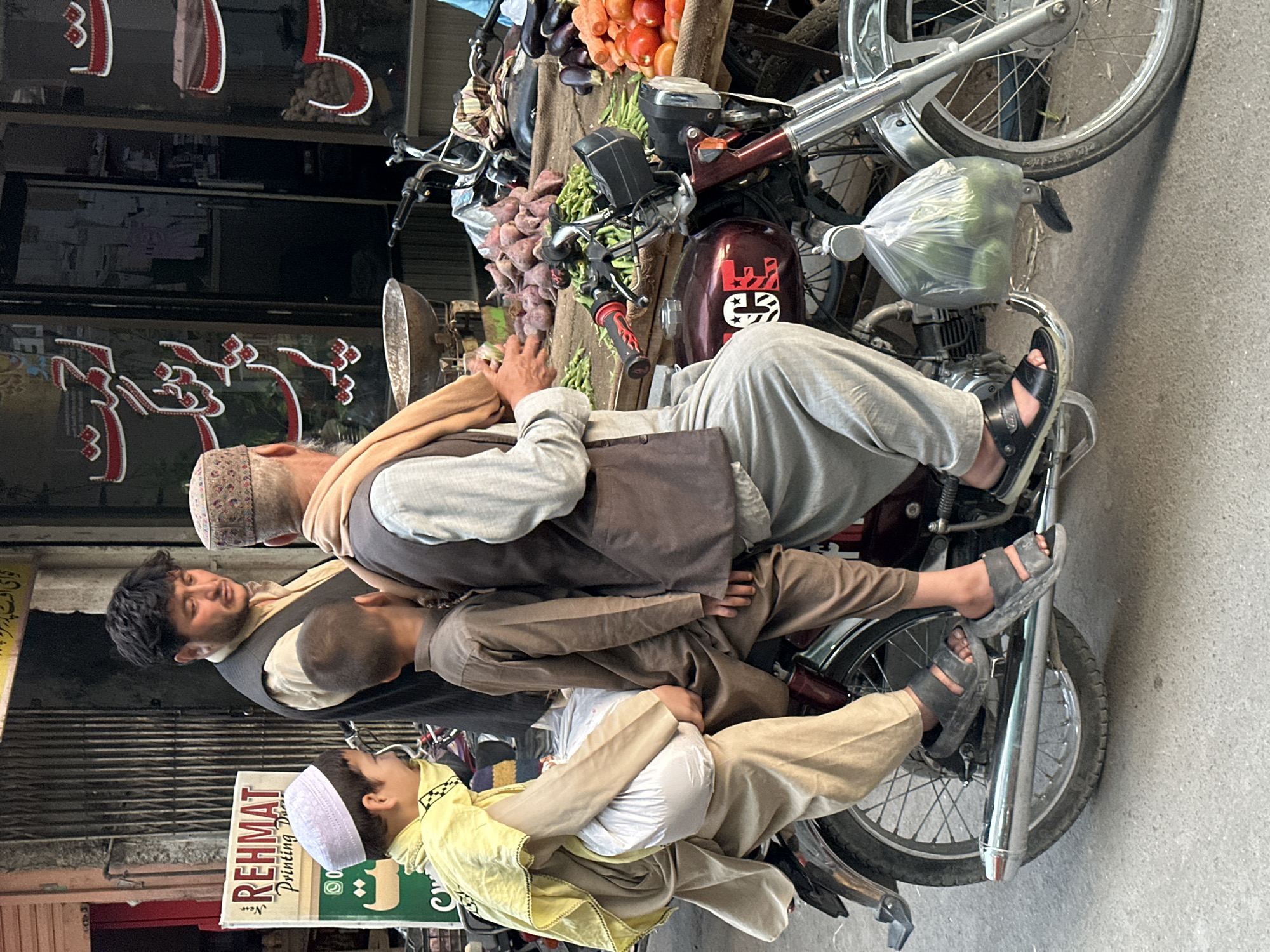 A color, medium shot depicts three people riding a motorcycle next to a storefront in Quetta City Tehsil, Pakistan. The man at the back of the motorcycle has gray hair and is wearing a brown cap, light-colored shirt, brown vest, gray pants, and black sandals. A young man is sandwiched between the first man and the young boy riding in the front. The young boy is wearing a white cap, white shirt, and a yellow jacket. There is a motorcycle in front of the one the three people are riding and there are fruits and vegetables displayed on it. There is a sign that says "REHMAT Printing" on the left side of the image.
