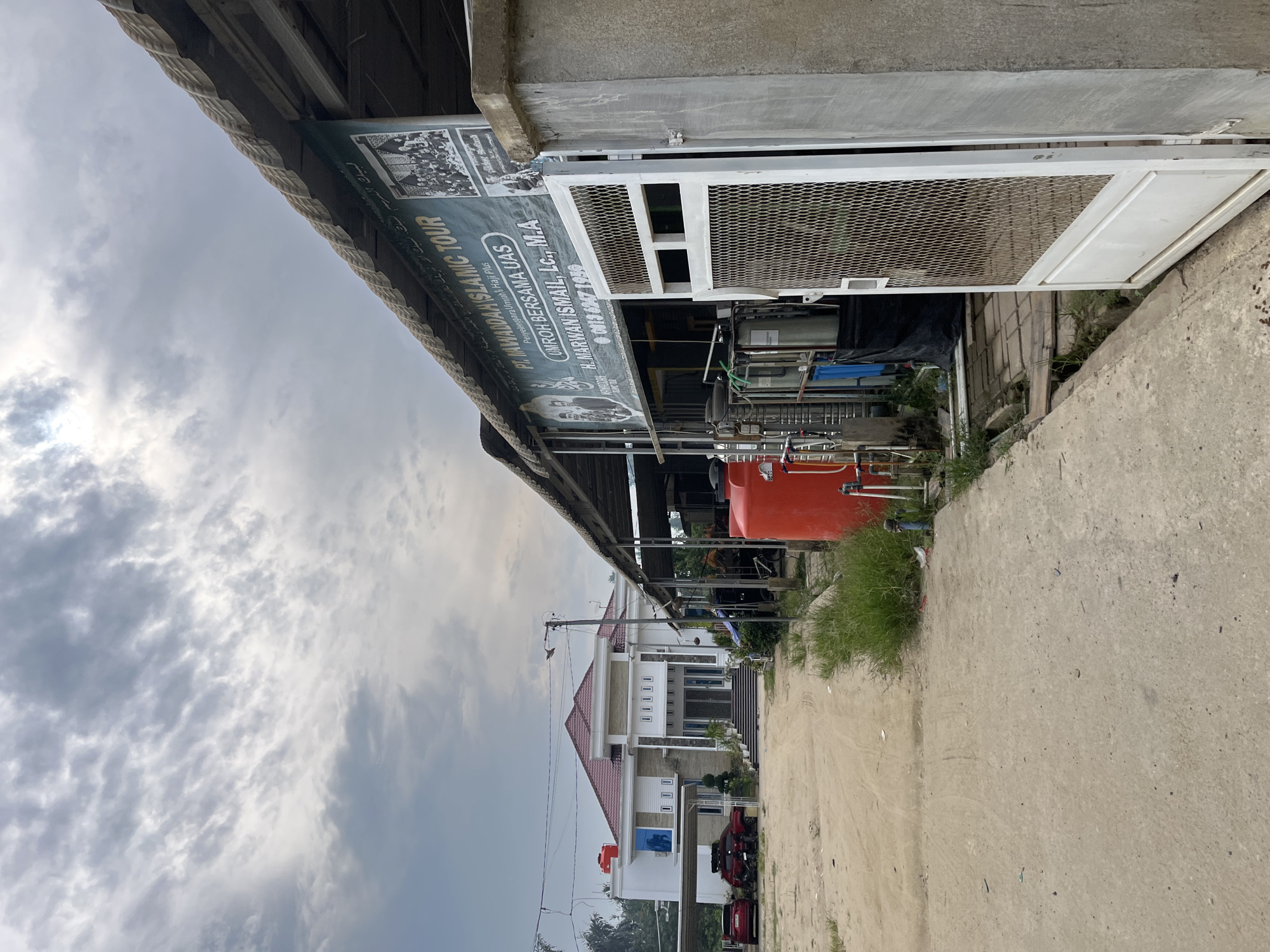 The image shows an outdoor scene in Tanahterban, Indonesia, with a modern white building with a red roof on the left, and a covered outdoor area with industrial equipment on the right. A dusty dirt road leads towards the building, with two red cars and one black SUV parked in front of it. The building features a grand entrance with stairs and decorative columns. To the right, a green banner advertising "PT. NAWADAH ISLAMIC TOUR" is visible, along with images and contact information. Industrial tanks, pipes, and other machinery are present under the covered area, with a large red water tank prominently displayed. Overcast skies with visible clouds dominate the upper portion of the image. The overall environment appears to be a mix of residential and commercial, with a slightly unkempt appearance in the foreground.