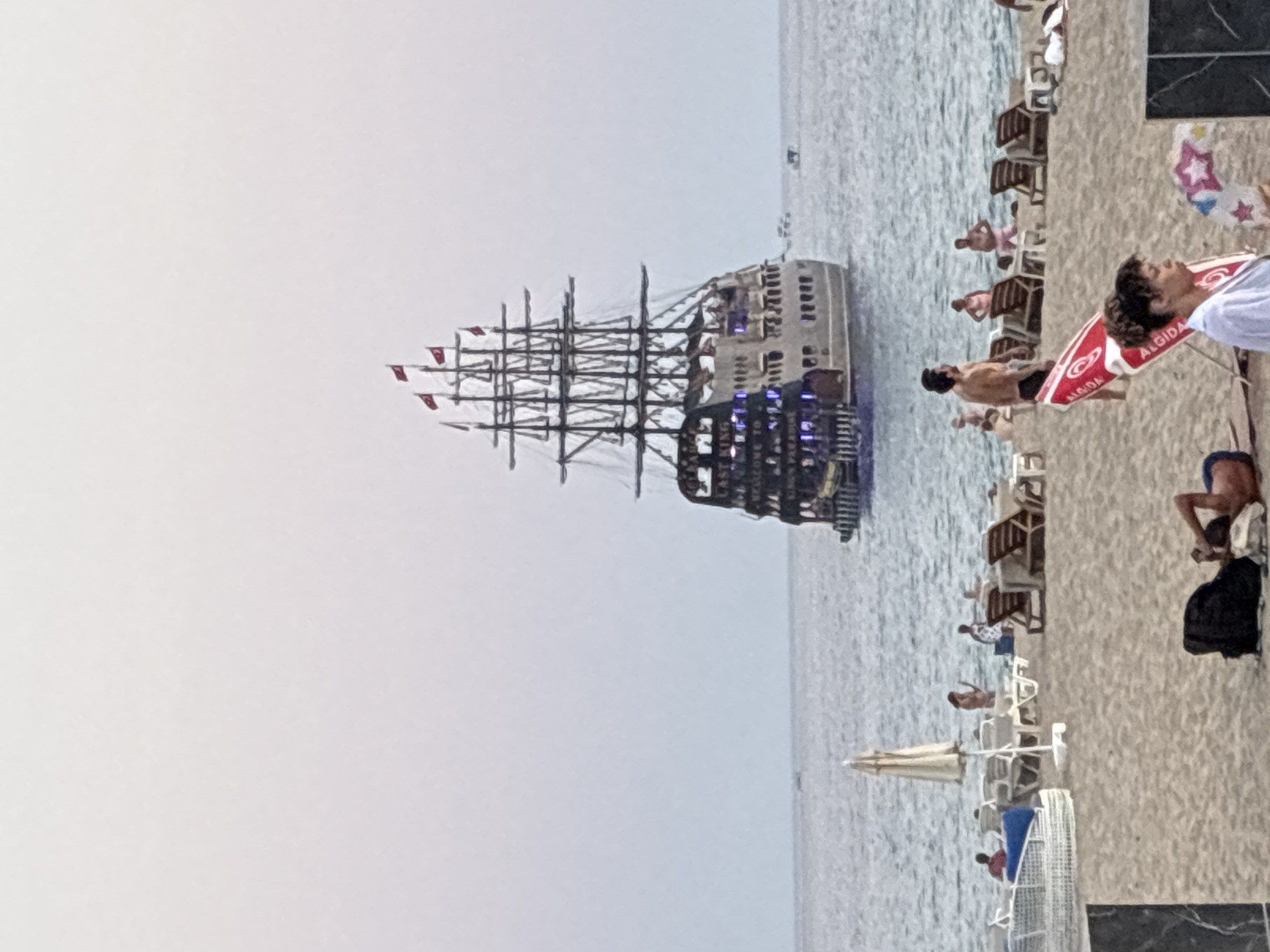 The image depicts a beach scene with a prominent sailing ship in the background, likely moored near the coast of Alanya, Turkey. The ship, a large galleon-style vessel, is illuminated with blue lights and displays text that reads "EAST KING WELCOME TO PARADISE". Several red flags are visible on its masts. The foreground shows a sandy beach with beach chairs, umbrellas, and several individuals. A man is lying on the sand, and others are visible in the background near the water and on the beach. The sea is calm, and the sky is overcast with a light, pale hue. The overall atmosphere suggests a leisurely day at a tourist destination.