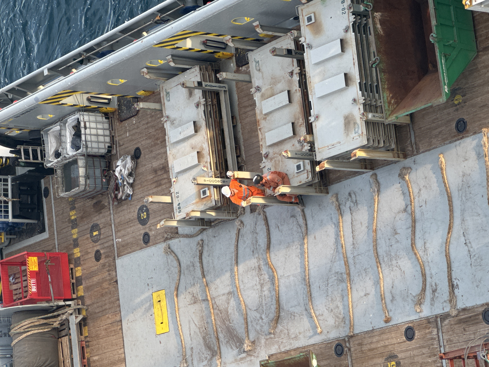 The image shows an aerial view of a ship's deck with two individuals working. The deck is made of wooden planks and concrete. Several large, white, metal containers or modules are stacked on the left side of the frame. To the right, there are several thick ropes lying on the concrete surface.
Two people, wearing orange jumpsuits and hard hats, are the primary subjects. One person is leaning on a metal structure, while the other is crouched down, seemingly inspecting or working on something near the base of the modules. The overall scene appears to be a work environment, likely on a vessel at sea, given the presence of the water in the background.
There are several visible labels and markings on the objects in the image. The white modules have stenciled text that includes "504-02" and "R-00002N". A red lifting cage on the far left has the text "BUTTI" on it. Yellow markings and black circles are also visible on the deck. The lighting suggests it is daytime, possibly late afternoon or early morning given the shadows. The weather appears calm, with no visible signs of rain or strong winds. The overall mood is one of industrial activity.