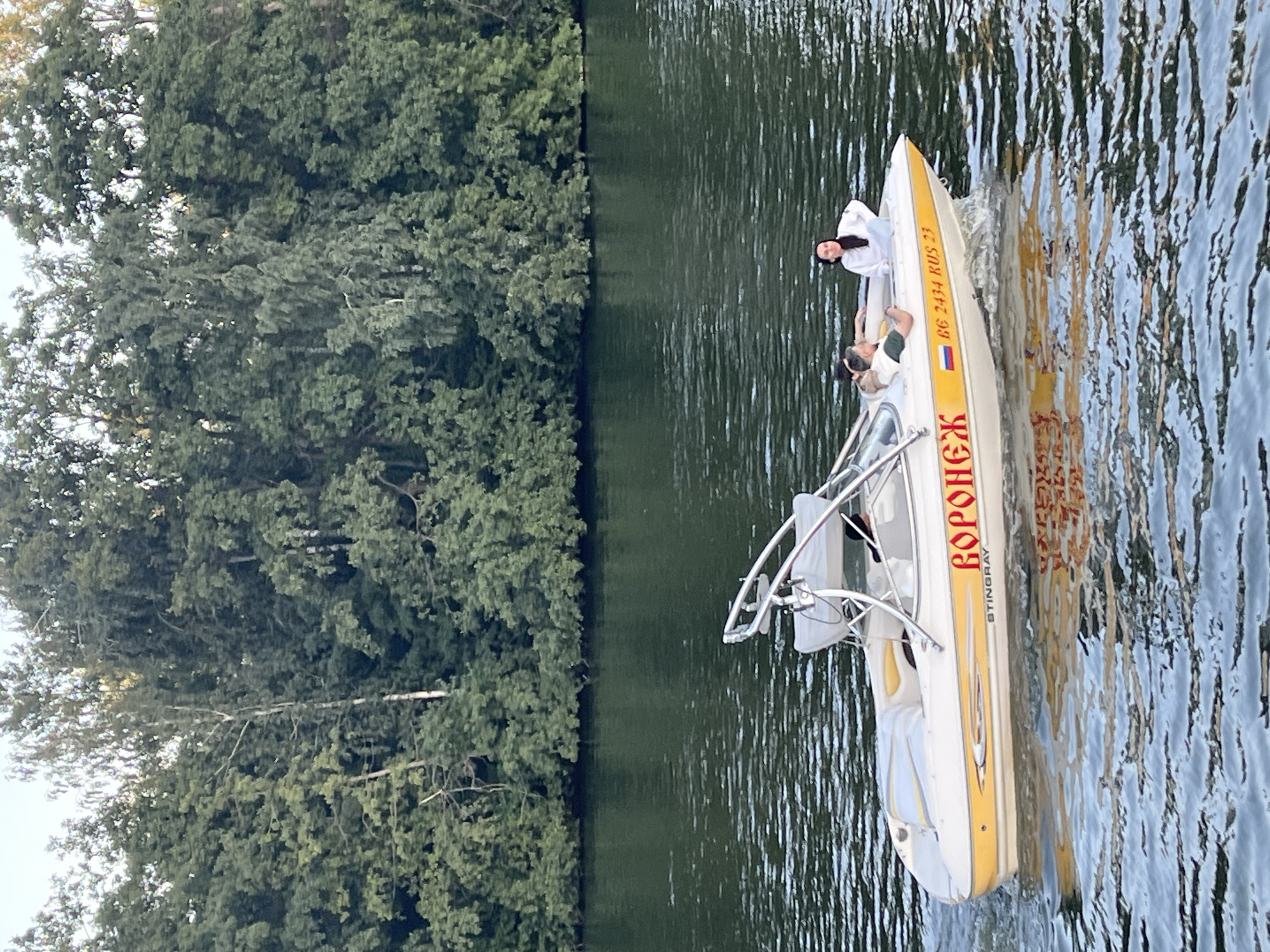 A white and yellow motorboat with a silver frame is moving on dark water. The boat has red Cyrillic lettering that reads "ВОРОНЕЖ" (Voronezh) and also displays "STINGRAY" and "BG 2434 RUS 23". Three people are on board. A man in a green shirt is seated, and a woman in a white hooded sweatshirt is standing. Another person, partially visible, is seated behind the man. The boat is creating a wake. In the background, a dense line of lush green trees lines the shore of a river or lake, with the sky visible above. The scene appears to be set in Novaya Usman, Russia, given the boat's markings.