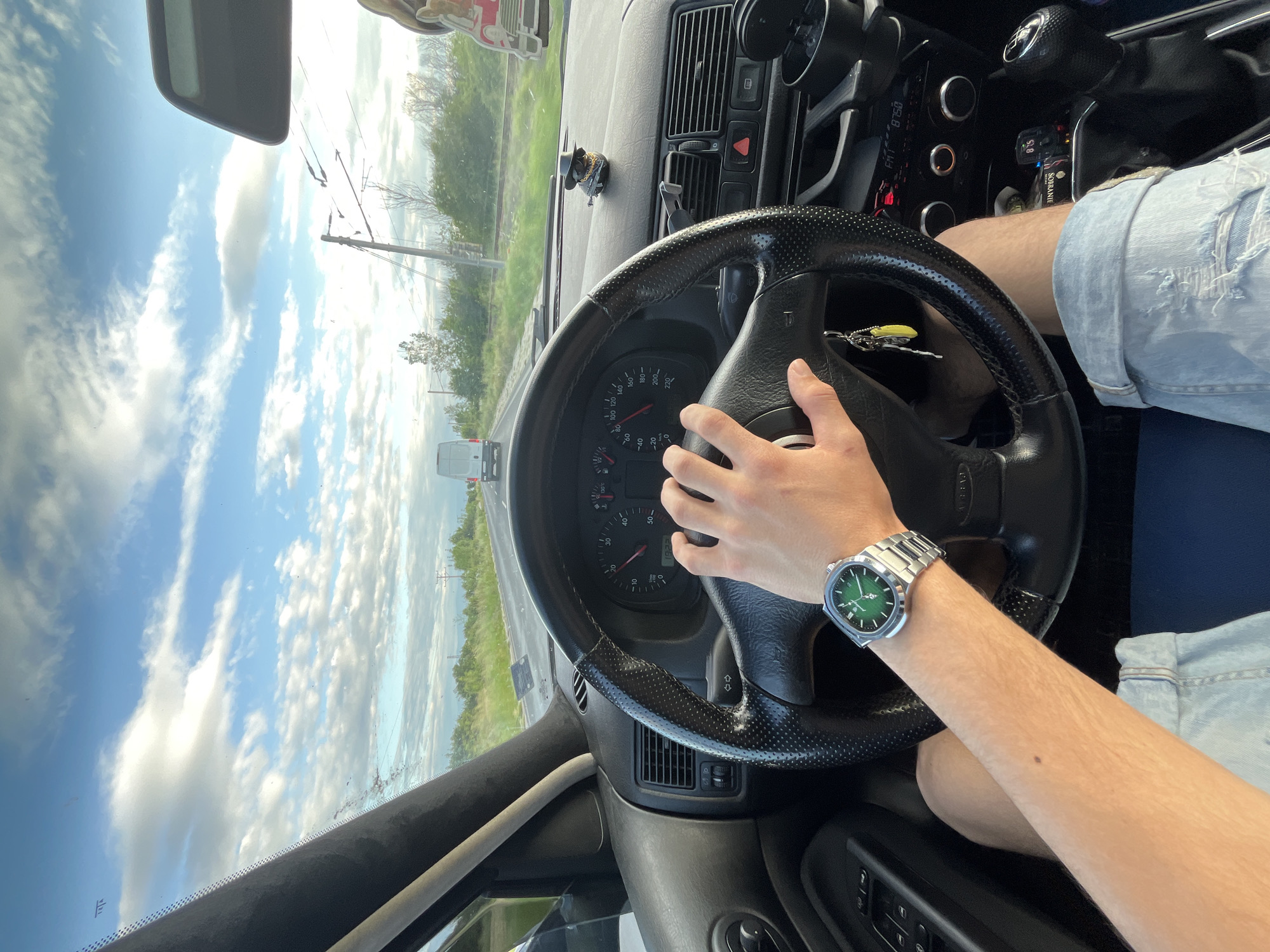 The image shows a person driving a car. The driver is holding the steering wheel with their left hand, wearing a silver watch with a green face on their left wrist. They are wearing light blue denim shorts. The interior of the car is visible, including the dashboard, speedometer, and other controls. Outside the car, there is a road, trees, and a cloudy sky. The location is the city of Dalgo Pole, Bulgaria.