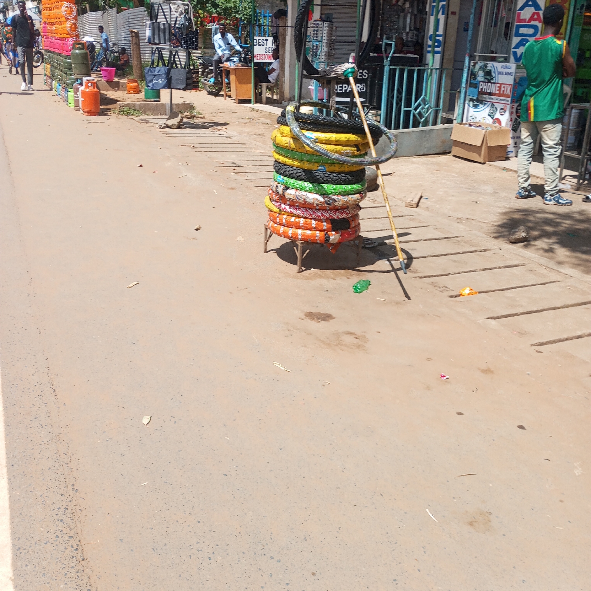 The image captures a street scene in Sheywe ward, Kenya. A stack of tires, various colors and patterns, sits on a small, makeshift wooden stand on the sidewalk. A long, wooden pole leans against the stack. Behind the tires, shops are visible, some displaying signs like "PHONE FIX" and "BEST REPAIR MEDSTOWN." A person in a green and yellow shirt stands near the shops. Other individuals are seen walking or sitting near storefronts in the background. Gas cylinders are stacked against a building in the midground. The road is cracked and shows signs of wear. The overall environment suggests a busy, commercial area.
