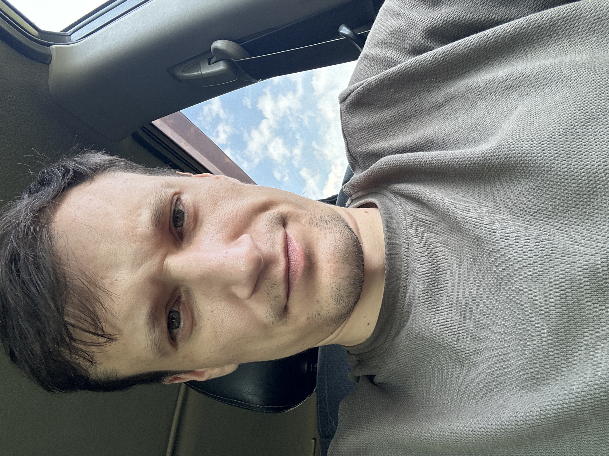 A close-up selfie shows a man sitting in the driver's seat of a car. The man has dark hair and light skin, and is wearing a brown t-shirt. He is looking directly at the camera with a neutral expression. Through the car window, a blue sky with white clouds is visible. The interior of the car, including the dashboard and a seatbelt, can be seen in the background. The man's facial features are the primary focus of the image. No specific location in Makiv, Ukraine is identifiable.