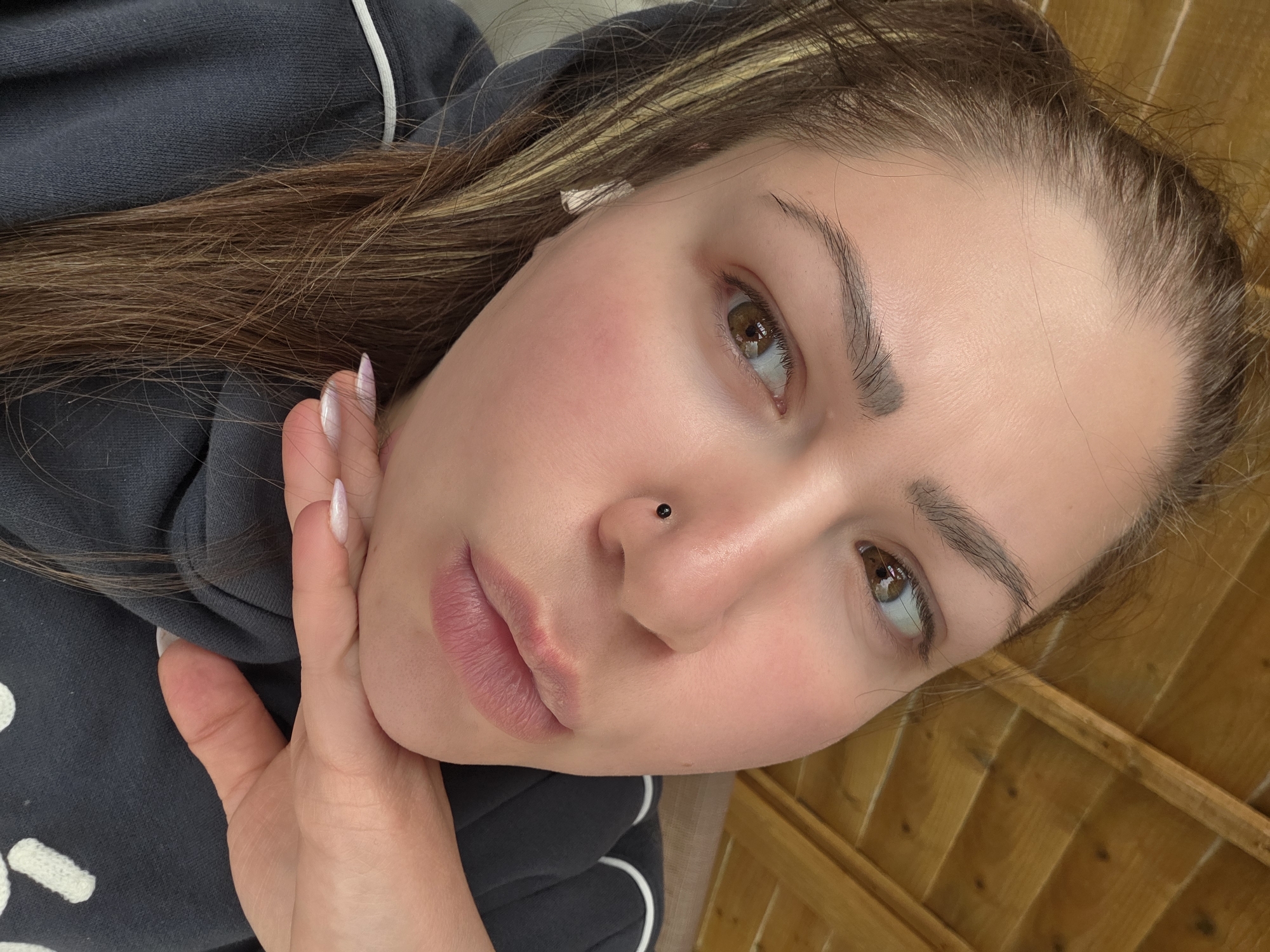 A close-up, angled portrait features a young woman, likely in her late teens or early twenties, looking upwards and slightly to the right. She has long, straight brown hair with subtle lighter highlights, cascading around her shoulders. Her brown eyes, with visible reflections of light, are framed by well-groomed eyebrows. A small, black stud piercing is visible on her right nostril. Her skin appears clear, with a natural flush on her cheeks, and her naturally pink lips are slightly parted.
She is wearing a dark grey hooded sweatshirt accented with distinct white piping details. Her left hand is gently placed against her jawline, showcasing long, almond-shaped fingernails painted with a light, possibly iridescent white polish.
The background behind her is composed of a warm-toned, wooden slatted structure, suggesting an outdoor setting such as a fence or decorative wall. The lighting appears soft and natural, indicating it is daytime. The image captures a casual, intimate moment, possibly a selfie. There is no visible text in the media file.