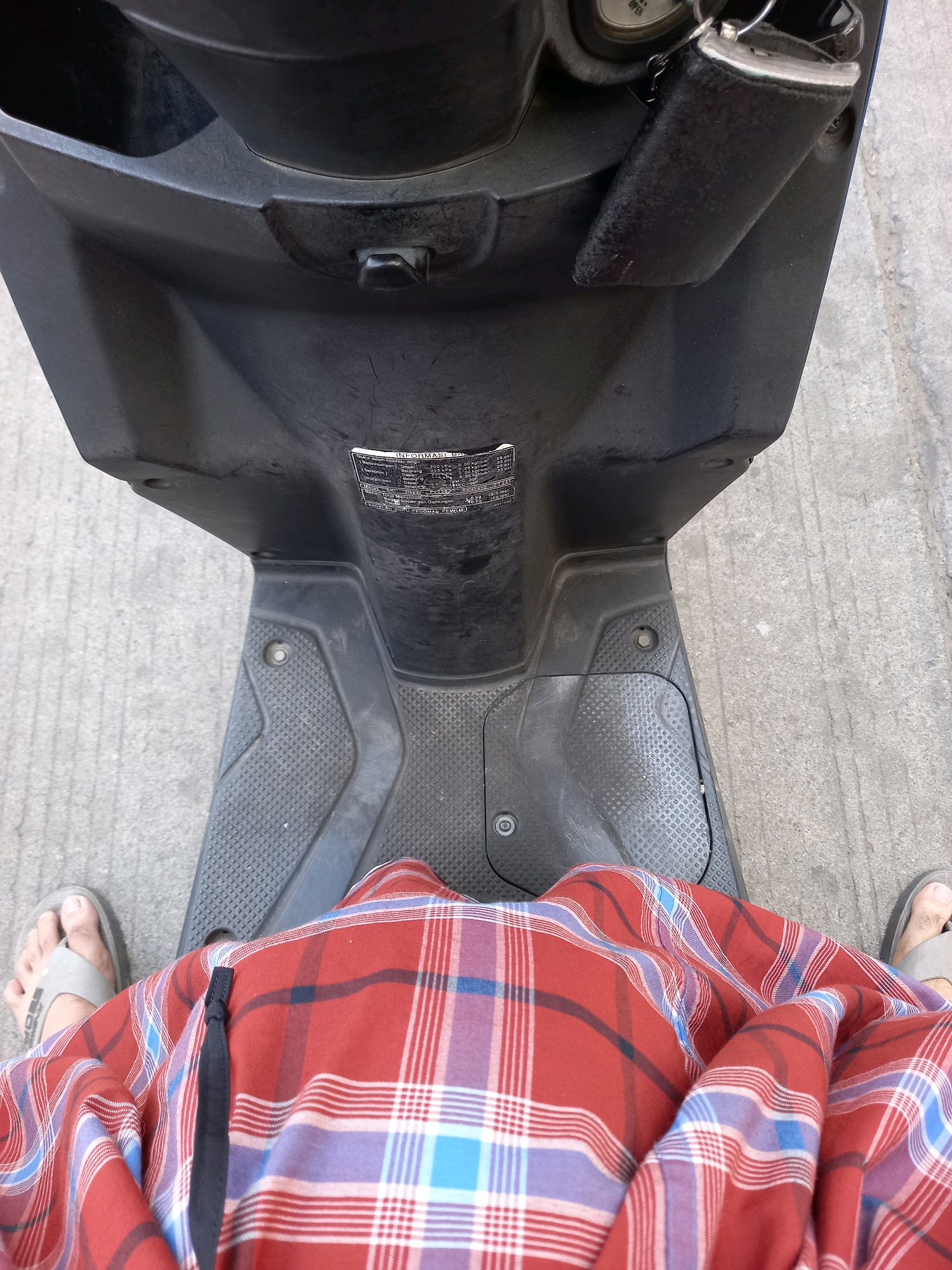 The image shows a first-person perspective of someone sitting on a black scooter. The person is wearing a red, white, and blue plaid sarong and grey sandals. The scooter's floorboard and leg shield are visible, as is a small compartment near the handlebars with a black key fob. The scooter is parked on a grey concrete surface, likely a street or sidewalk in the city of Samarinda, Indonesia. The lighting suggests it is daytime.