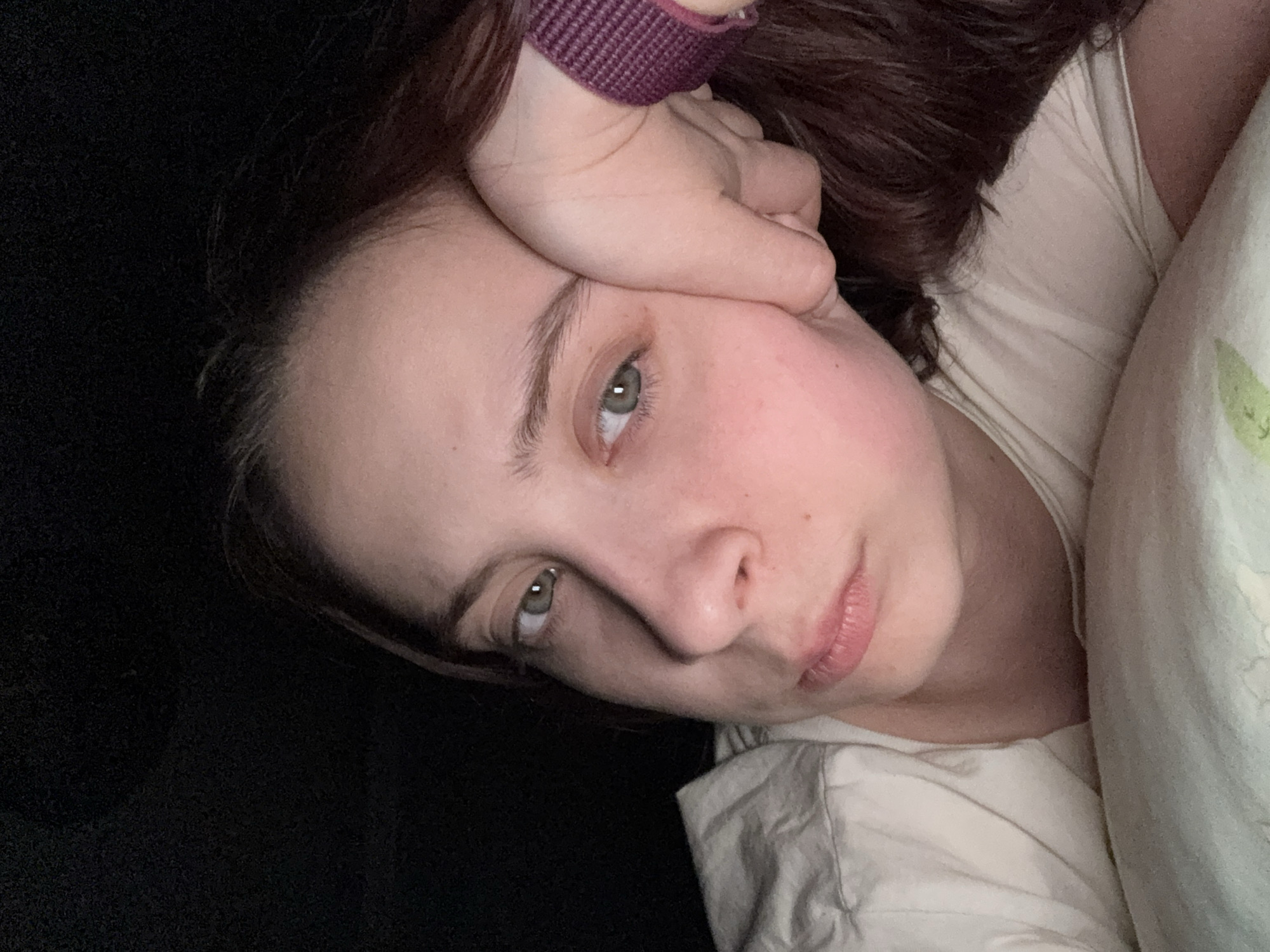 A close-up, tilted indoor portrait of a woman with fair skin, light eyes, and dark hair. She is lying down, with her head resting on her hand, which is adorned with a purple wristband. She is wearing a light-colored shirt and resting against a pillow with a subtle leaf pattern. The background is dark, contrasting with the light complexion of the woman. The location is unknown and there is no indication of Belaya Pashnya, Russia.