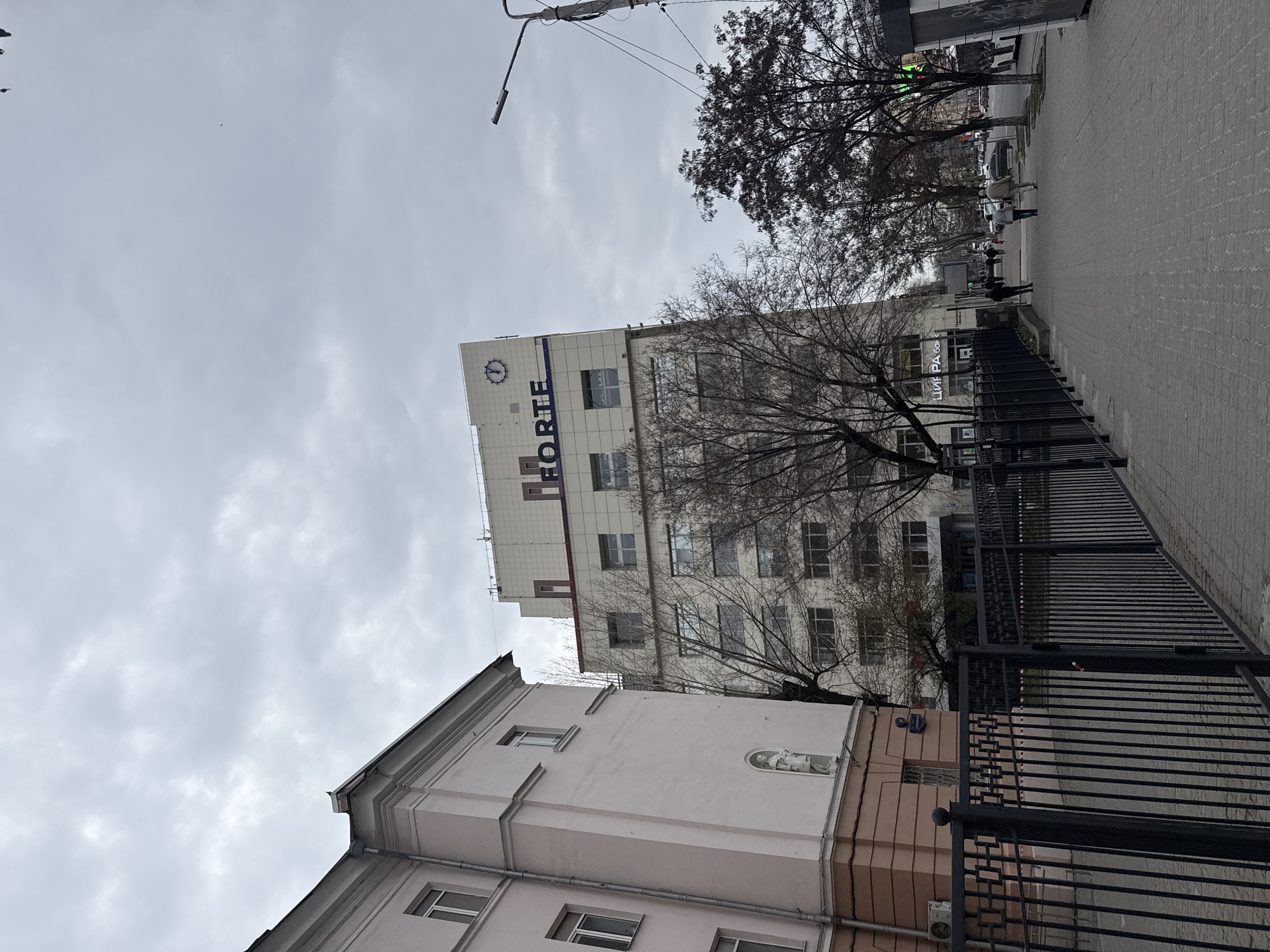 The image depicts a street scene in Rostov-on-Don, Russia. A tall, multi-story white building with blue lettering "FORTE" is visible, along with a clock. A pink building stands adjacent to it. Bare trees partially obscure the view of both structures. A black metal fence lines the sidewalk. Pedestrians are walking on the grey-paved sidewalk. The sky is overcast with grey clouds. The street also has lighting fixtures.