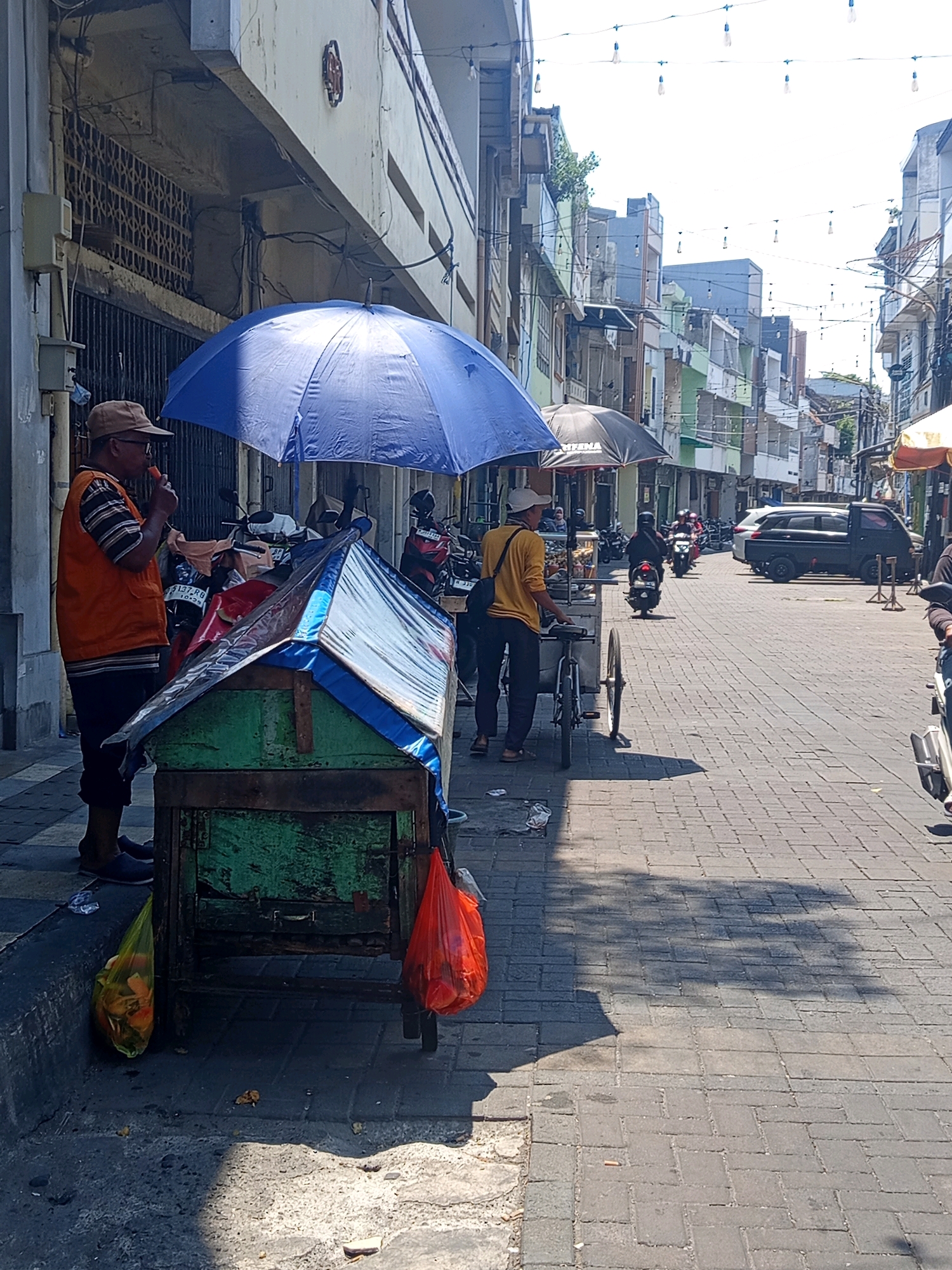 A street scene in Semarang, Indonesia, features a man in an orange vest and a brown hat standing next to a green cart covered with a blue and clear tarp. He is holding and eating an orange-colored food item. Another man, wearing a yellow shirt and a white cap, stands by a bicycle cart. In the background, motorcycles and a black van are parked on a brick-paved street lined with multi-story buildings. A blue umbrella shades the area near the green cart, and a black umbrella is further down the street. Orange and yellow plastic bags are visible on the ground near the cart, and a shadow of a tree is cast on the pavement. The sky is bright and clear.