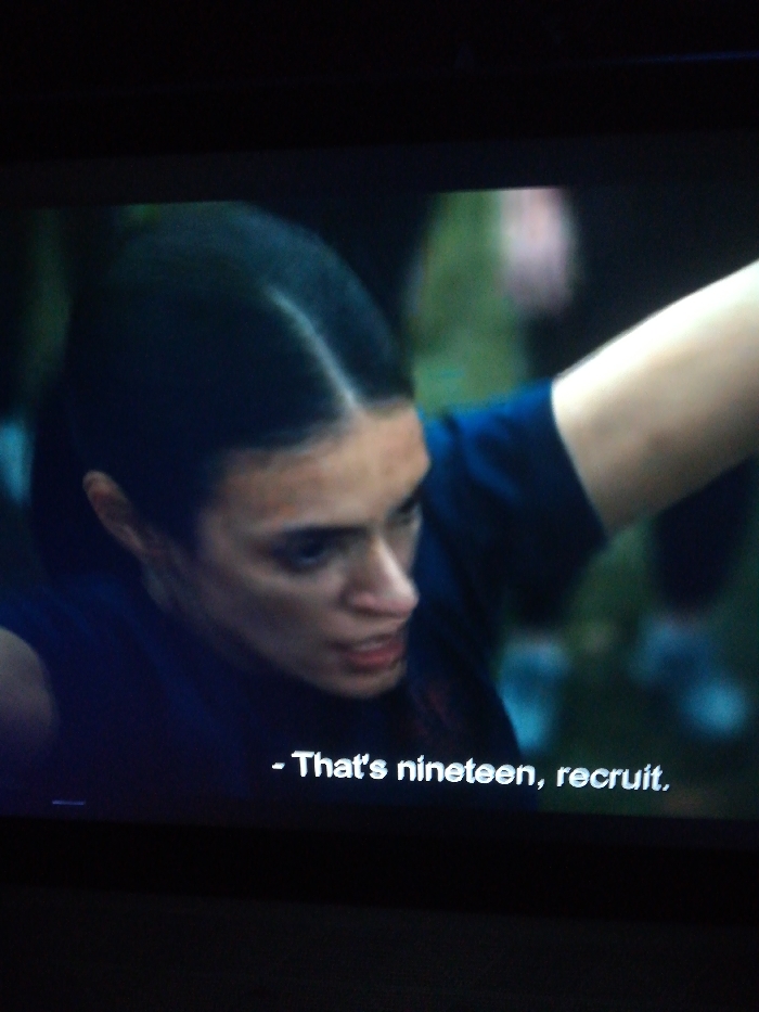 A frame captures a woman with dark hair pulled back from her face, her expression focused and serious. The backdrop appears to be an outdoor setting with other individuals blurred in the background. She is wearing a dark blue shirt. Text overlay at the bottom of the image reads, "That's nineteen, recruit." The lighting in the scene is somewhat dim, casting shadows on the woman's face and emphasizing her determined expression.
