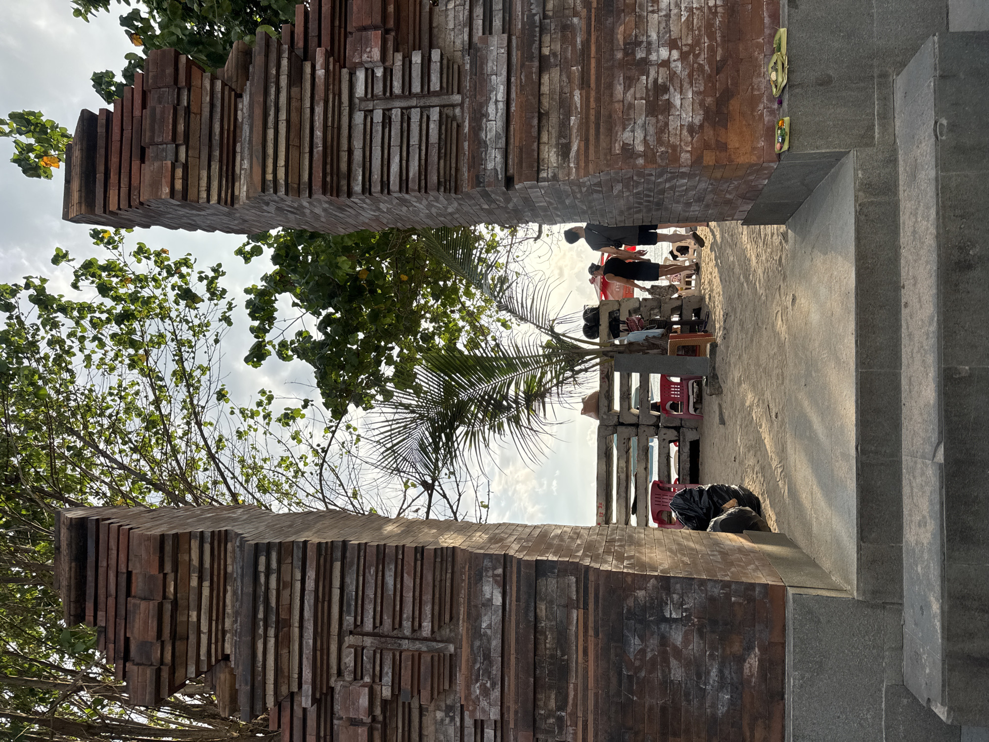 This image depicts a view through a brick archway in Kuta, Indonesia. The archway, composed of stacked bricks, frames a scene of a sandy area with trees and people. On the right side of the archway are stone steps. The people in the distance appear to be standing and sitting. There's a black plastic bag near the stairs.