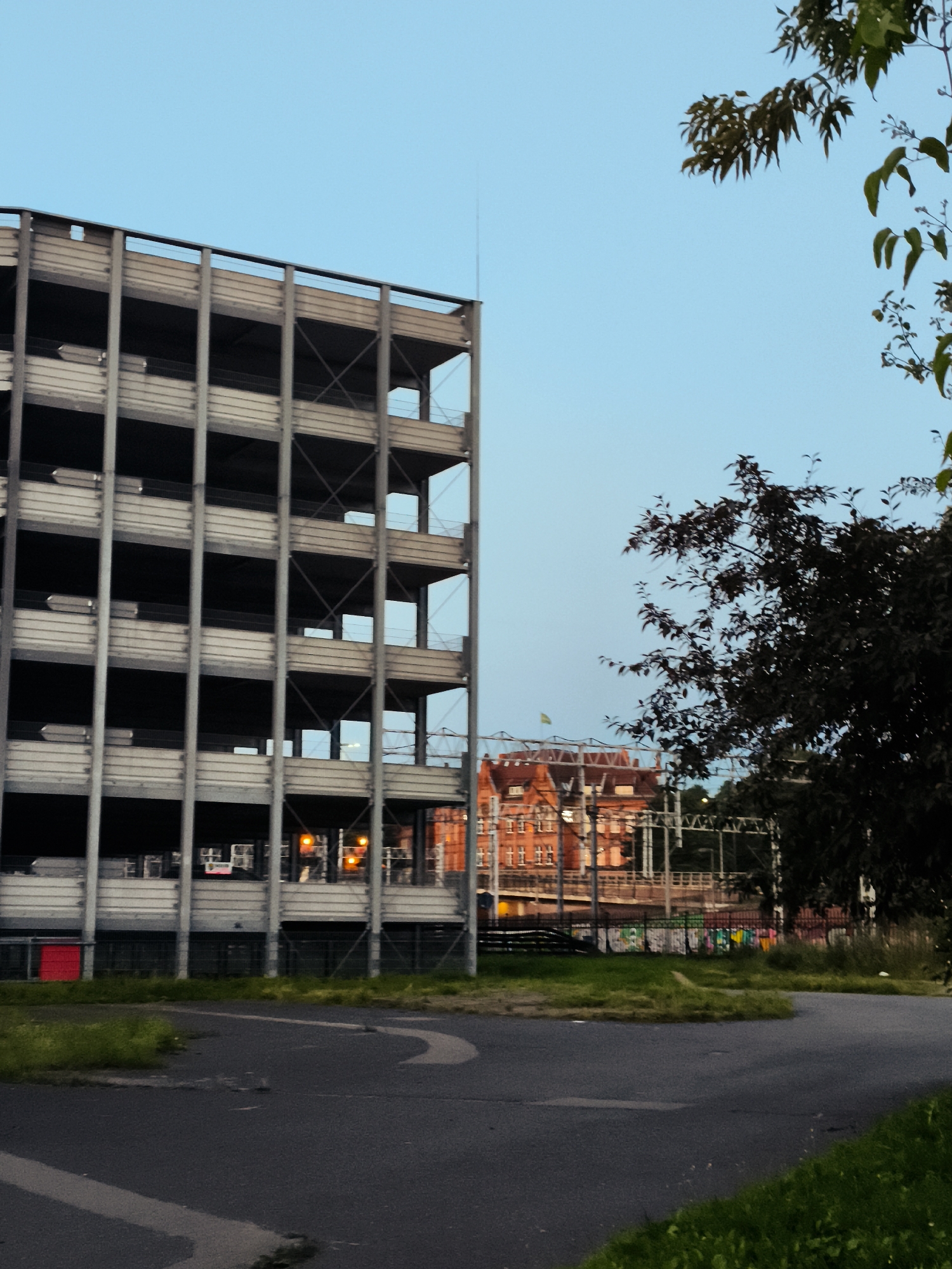 The image depicts an exterior view of a multi-level concrete parking garage with a grey metal frame, viewed from a paved area with some grass. The garage has open-air levels, with visible supports and diagonal bracing. On the upper levels, orange lights are illuminated, suggesting activity within. In the background, a red brick building with multiple windows and a pitched roof is visible. Trees with dark green foliage are also present, partially obscuring the background. The sky is a clear, pale blue, indicating it is likely dusk or early morning. There are no people or animals visible in the image. The scene is located in the city of Gdańsk, Poland.
