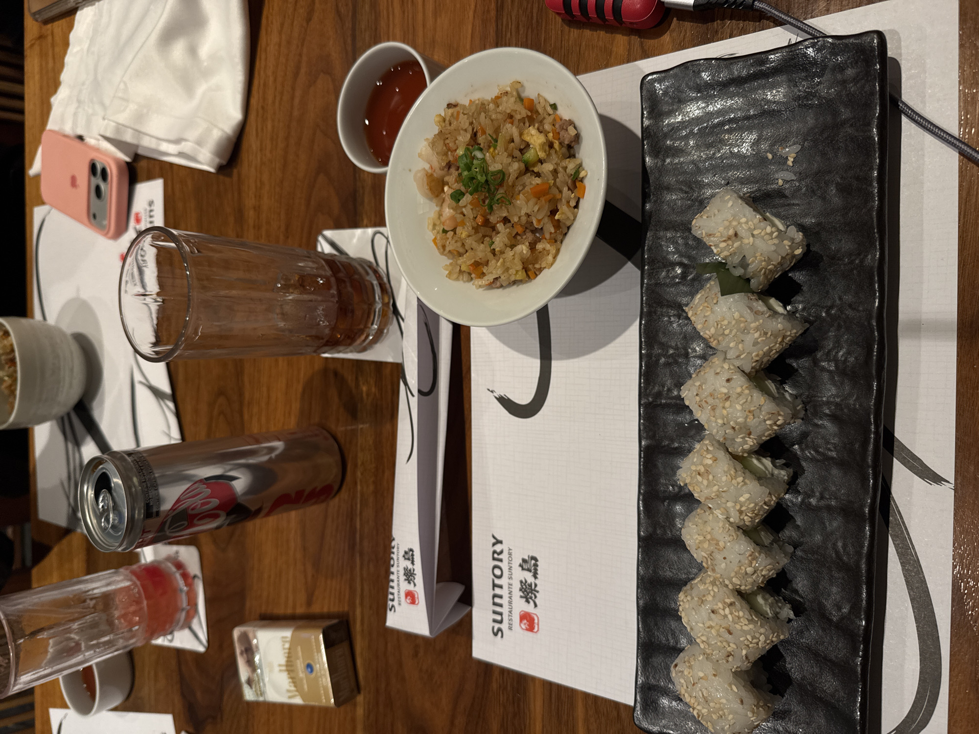 The image depicts a wooden table setting in what appears to be a Japanese restaurant. The items on the table include a plate of sushi rolls covered in sesame seeds, a bowl of fried rice with green onions, a small dish of dipping sauce, a glass of liquid, a can of Coca-Cola, a pack of Marlboro cigarettes, a mobile phone in a pink case, and a white cloth. Chopsticks in wrappers are also present, with the "Suntory Restaurante Suntory" logo printed on them. Based on the signage, this scene is likely from a Suntory restaurant, although identifying the specific location in Puebla, Mexico requires additional visual clues or information beyond the image.