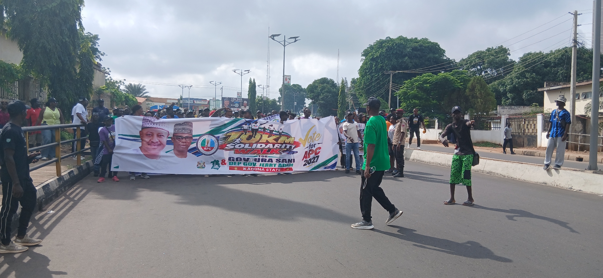 This image captures a street scene in Unguwar Shanu, Nigeria, where a large group of people are participating in a solidarity walk. A prominent banner, held by several individuals, features images of two men and text promoting a "10KM Solidarity Walk for Gov. Uba Sani & Dep. Gov. Jerry Adam, Kaduna State." The banner also includes the APC logo and the year 2027.
The participants are a mix of men and women, many dressed in casual clothing, while some are wearing t-shirts and caps associated with the political party. The walk appears to be taking place on a paved road, with trees and buildings lining the street. Several lampposts are visible in the background, along with utility poles and wires. Some individuals are walking alongside the banner, while others are gathered behind it. A few men are standing on a concrete barrier at the side of the road, observing the procession. The sky is overcast, suggesting a cloudy day. The overall atmosphere suggests a political rally or campaign event.