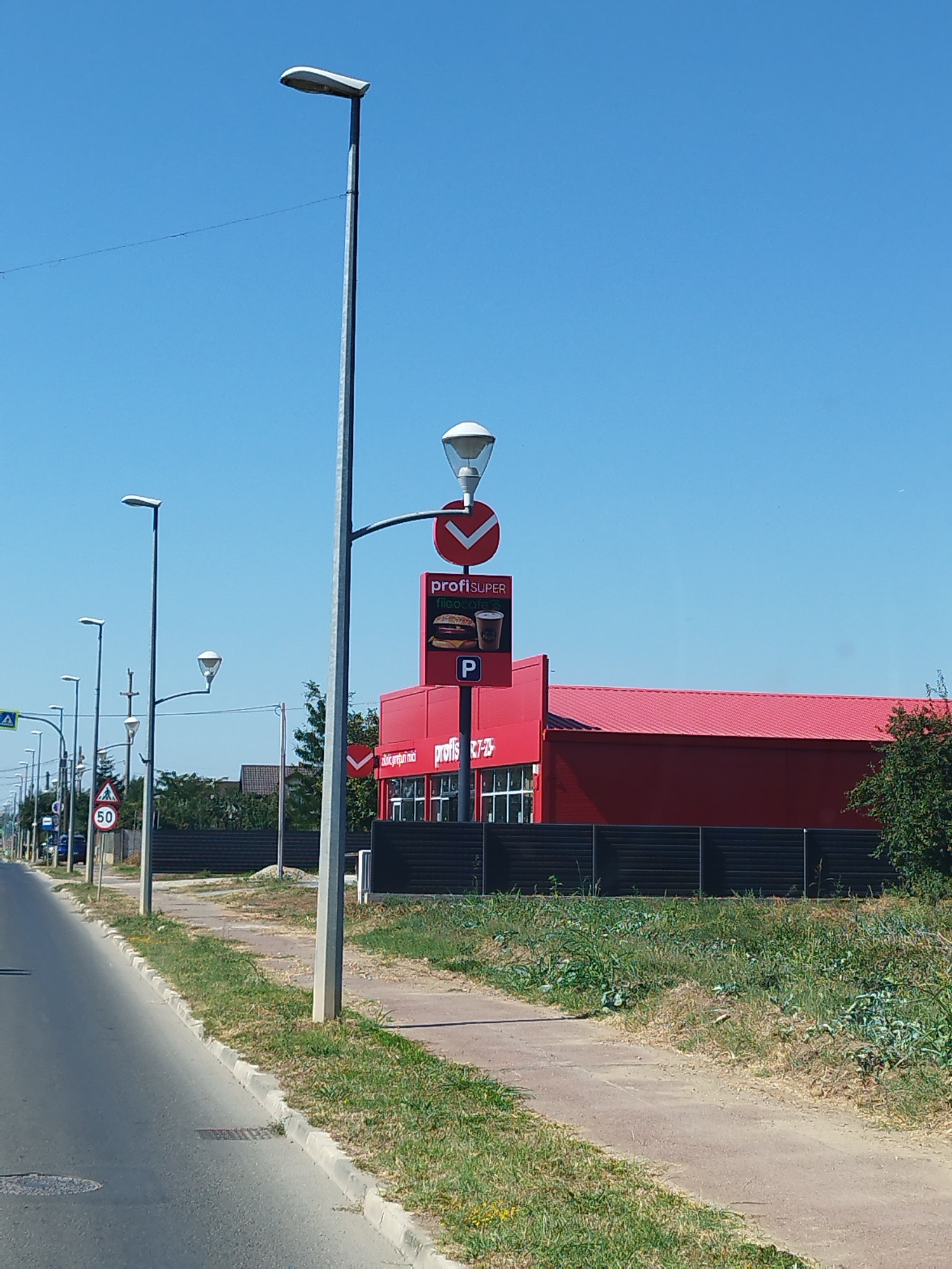 The image shows a bright, sunny day in Buftea, Romania. A red building with a dark fence in front of it stands on the right side of the frame. The building has a sign that reads "profiSUPER". On the left side of the image, there is a road with a sidewalk. Several streetlights are visible along the sidewalk. A speed limit sign indicating "50" is also present. The sky is clear and blue. The scene appears to be a commercial area with a grocery store or supermarket.