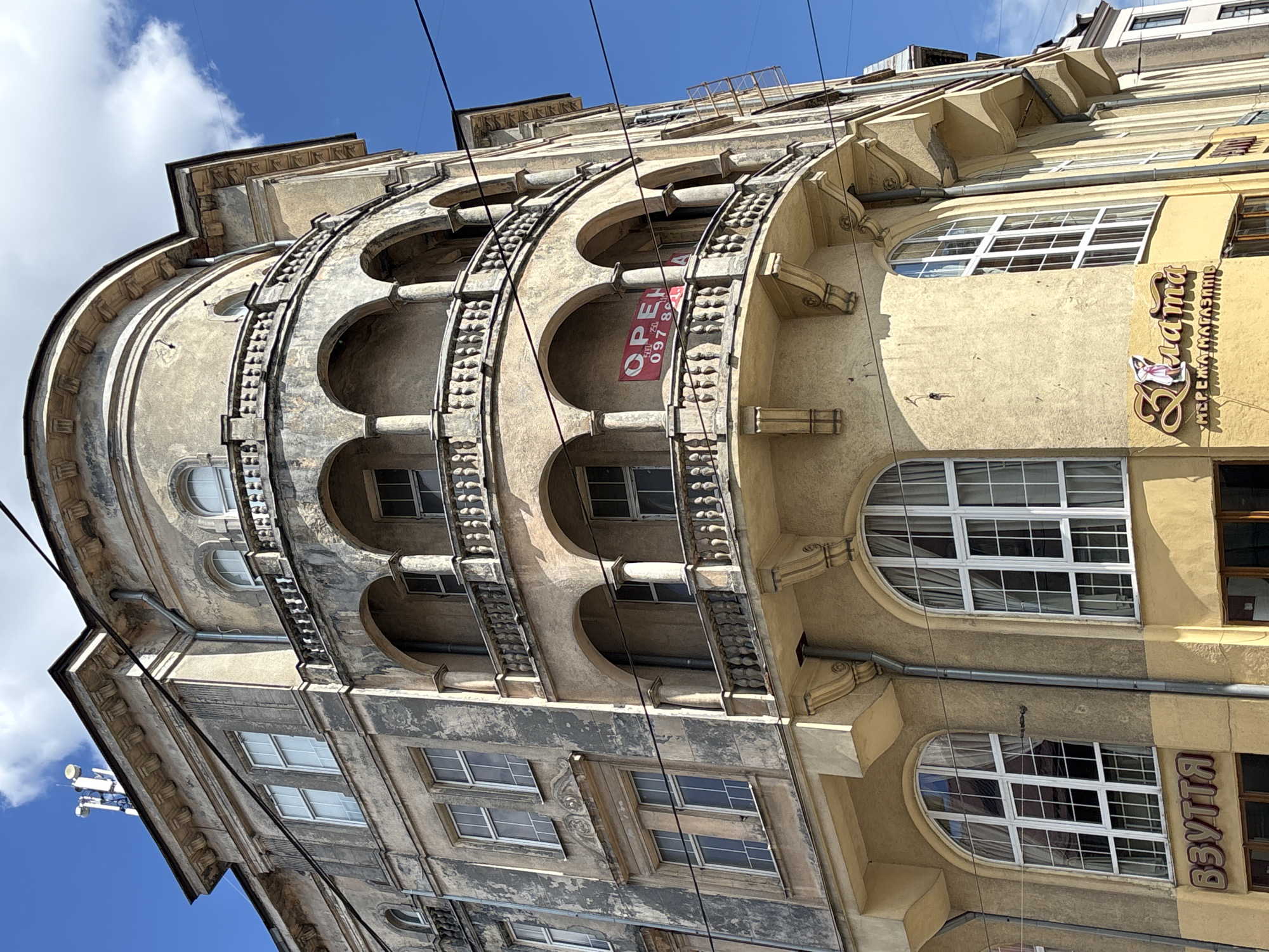 The image shows the exterior of a building in Lviv, Ukraine. The building has an ornate facade with a rounded section featuring arched openings supported by columns. A sign with the word "OPEN" is visible in one of the arches. The building is multi-storied, with windows on each level. The facade is a mix of beige and gray tones, showing signs of age and weathering. Above the rounded section, there are more stories with rectangular windows. Power lines are visible against a blue sky with some clouds. The right side of the building has a storefront with a sign in Ukrainian.