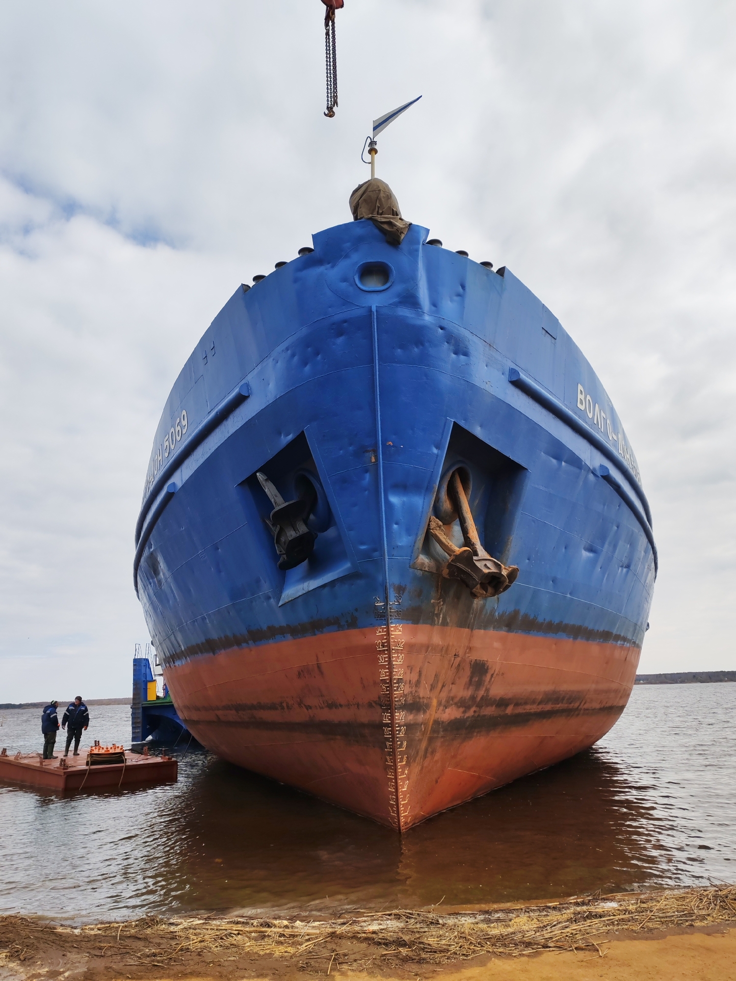 A large cargo ship, painted blue above the waterline and red below, is prominently featured from a head-on perspective, partially out of the water on a muddy bank. Its V-shaped bow points towards the viewer, revealing two large anchors stowed in their hawseholes on either side. Visible white draft marks run vertically on the bow, indicating depth measurements, with the current waterline appearing around the 22-24 mark.
Text painted on the port (left) side of the ship's bow reads "ВОЛГО-ДОН 5069" (Volgo-Don 5069), while "ВОЛГО-ДОН" (Volgo-Don) is also visible on the starboard (right) side. Above the ship, a crane hook and chain are visible against the cloudy sky, suggesting ongoing lifting or construction work.
To the left of the ship, two individuals dressed in practical workwear stand on a small, rectangular floating pontoon, observing the scene. The water around the pontoon and the ship is calm and appears brownish. The immediate bank is muddy, strewn with dried reeds and grass. The sky is overcast, indicating a cloudy daytime setting. This scene depicts a ship, likely a river-sea vessel, undergoing work at a shipyard or docking area in Rybinsk, Russia.