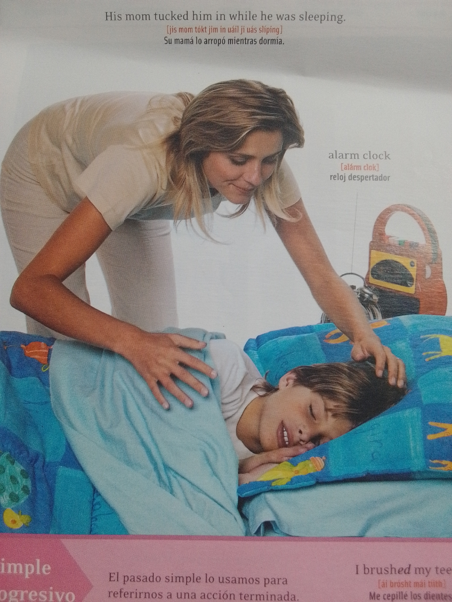 The media file depicts a heartwarming scene of a mother tucking her young son into bed. The boy, with a peaceful expression and eyes closed, is nestled under a light blue blanket adorned with colorful cartoon characters. His mother, with blonde hair and a gentle smile, leans over him, her hand resting tenderly on his head. The setting appears to be a bedroom, implied by the presence of a bed and the mother's actions. In the background, an alarm clock and a retro-style radio are visible on a bedside table, suggesting it might be morning or evening.
The visible text provides translations and explanations related to the scene. The English sentence "His mom tucked him in while he was sleeping" is accompanied by a phonetic transcription and a Spanish translation: "Su mamá lo arropó mientras dormía." Another section of text mentions "alarm clock" with its phonetic spelling and Spanish translation, "reloj despertador." Further down, there's text in Spanish discussing the use of the simple past tense ("El pasado simple lo usamos para referirnos a una acción terminada") and English text saying "I brushed my teeth" with its phonetic transcription and Spanish translation, "Me cepillé los dientes." This juxtaposition of languages and phrases suggests the media file might be part of an educational resource, possibly for language learning. The overall mood is one of warmth, care, and domestic tranquility.