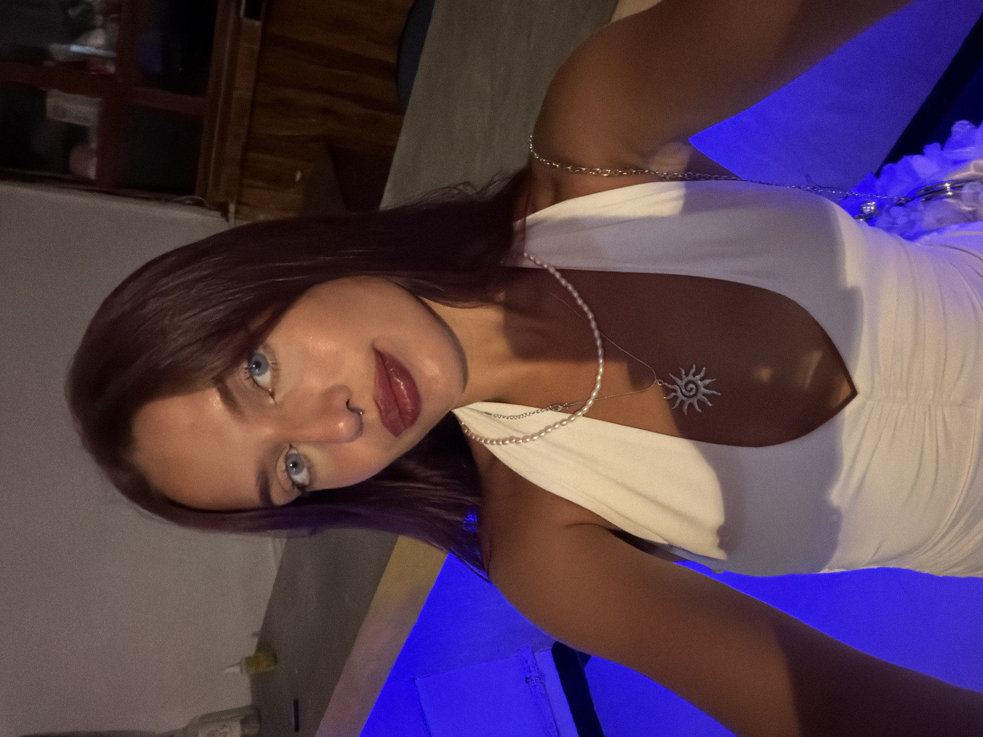 A young woman with dark red hair and light blue eyes is captured in a close-up selfie. She is wearing a white halter dress with a plunging neckline, accessorized with a pearl necklace and a sun pendant. A septum piercing is visible on her nose. The background is dimly lit, with a blue glow emanating from what appears to be a pool or water feature. In the background, there are wooden structures and shelves with various objects, suggesting an indoor setting perhaps in a bar or lounge. The overall ambiance suggests a nighttime social setting. The location is identified as Arugam Bay, Sri Lanka.