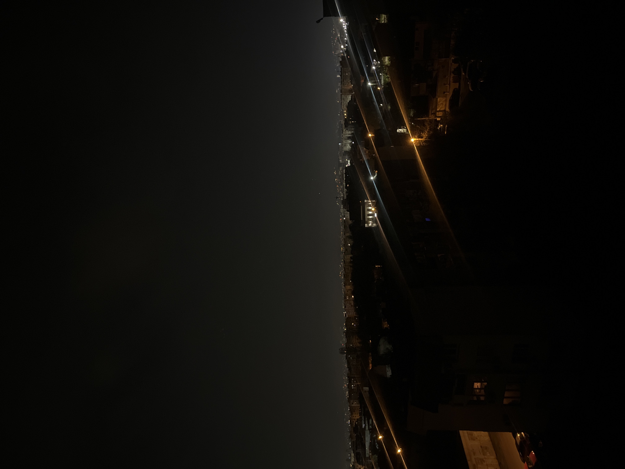 The image depicts a nighttime cityscape, possibly in Sofia, Bulgaria, based on architectural styles and urban layout. The scene is dominated by darkness, with artificial lights illuminating the area. The streetlights are visible, casting a yellowish glow on roads and buildings. There's a clear distinction between the brightly lit areas and the dark sky. The perspective is elevated, suggesting a view from a high-rise building. Buildings line the street and appear to be of varying heights. The composition is such that the cityscape stretches towards the horizon. There is no sign of people or other identifiable subjects.