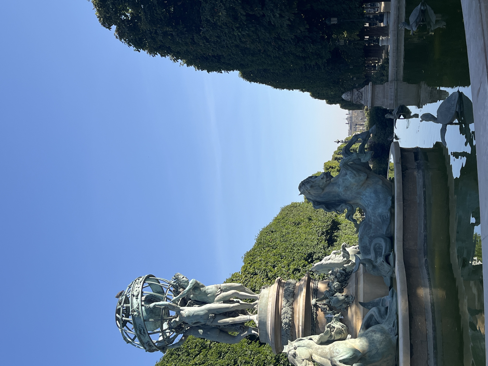 The image shows the Fountain of the Chariot of Apollo in the Tuileries Garden, Paris, France. The fountain features a large bronze sculpture of figures holding up a globe, with the lower part of the fountain depicting a chariot pulled by horses and other sea creatures. The sculpture is surrounded by a pool of water and manicured trees line the pathway behind it. The sky is a clear blue, and the sunlight casts shadows on the fountain and the water. There are several bronze sculptures of turtles in the water. In the background, a building with a dome is visible.