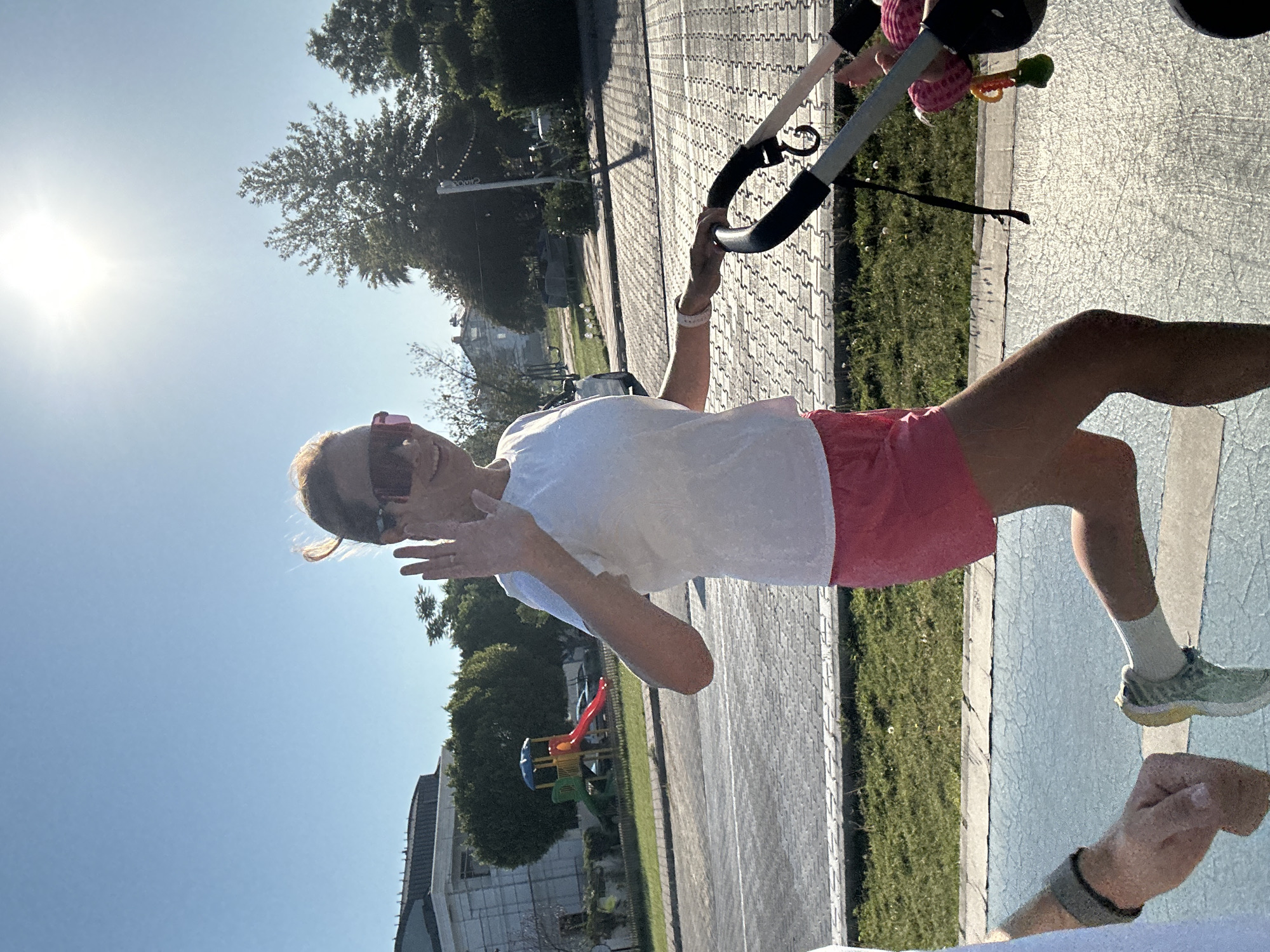 A woman, wearing a white t-shirt, pink shorts, sunglasses, and light-colored sneakers with white socks, is depicted in motion, pushing a dark-colored baby stroller. She is positioned slightly right of the frame's center, captured mid-stride with her left leg extended forward and bent at the knee, and her right foot partially visible behind. Her left arm is raised, hand open in a waving gesture, while her right hand grips the stroller handle. She is smiling, with the sun's glare reflecting off her sunglasses. A baby's feet and lower legs, clad in pink attire, are visible within the stroller on the right side of the frame, along with a colorful toy hanging from the stroller's frame.
To the left of the woman, a person's left arm and hand are visible in the foreground, wearing a dark wristband. The ground beneath them is a light blue paved surface, possibly a dedicated pathway, which transitions to a grey-patterned paved sidewalk further back. A strip of green grass separates the sidewalk from a paved road.
In the background, a children's playground with a red slide and blue and yellow structures is visible behind a fence. Beyond the playground, a large white building with dark roofs and scaffolding on one side stands against a clear, bright blue sky. Numerous green trees and shrubs are scattered throughout the background, indicative of a sunny, outdoor environment. The sun is high and bright, positioned in the upper right quadrant of the sky, casting distinct shadows. The architecture and vegetation are consistent with a Mediterranean urban setting, specified as the city of Fethiye, Turkey.