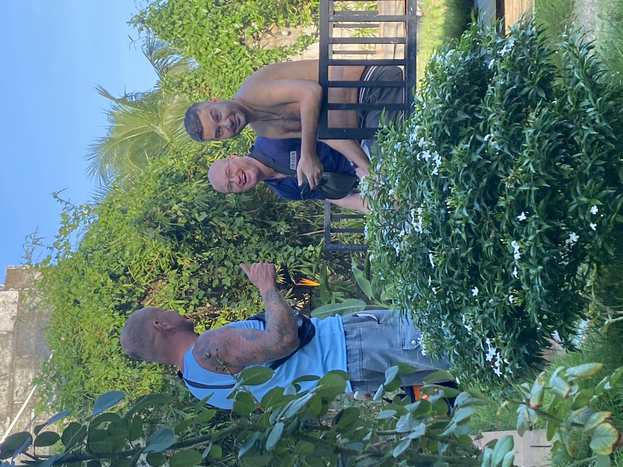 Three men are visible outdoors, with lush green foliage and a bright blue sky. The man on the left, wearing a light blue tank top and grey cargo shorts, stands with his back partially to the camera, giving a thumbs-up. He has tattoos on his left arm and a black bag slung across his chest.
Two men are seated on a dark metal bench to the right. The man in the middle is wearing a dark blue polo shirt with a "PRADA" logo and is holding a phone. He is laughing. The man on the right is shirtless, wearing dark shorts with a white stripe. He is looking towards the camera with a slight smile.
In the foreground, a dense green bush with small white flowers is prominent, partially obscuring the view of the men. The environment suggests a tropical or subtropical setting, likely in Mũi Né, Vietnam, known for its beaches and green landscapes. The lighting indicates it is daytime, with the sun casting shadows.