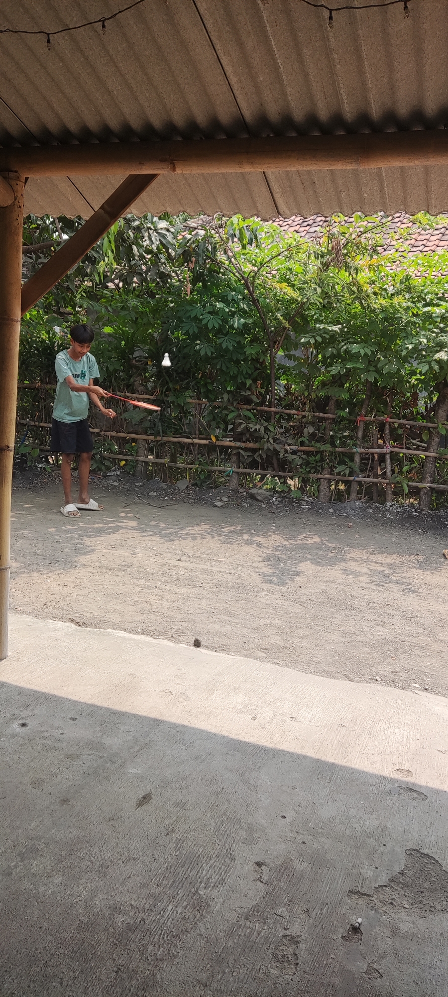 A young Indonesian boy is playing badminton in an outdoor area in Karangwangun, Indonesia. He is wearing a light blue t-shirt and dark shorts, with white sandals on his feet. He is holding a red badminton racket and is in the process of hitting a white shuttlecock, which is in mid-air. The background consists of lush green foliage and a rustic bamboo fence. The ground is a mixture of dirt and small stones. A corrugated roof structure extends from the left side of the frame, providing shade. The scene appears to be a casual outdoor setting, possibly a backyard or a local recreation area.