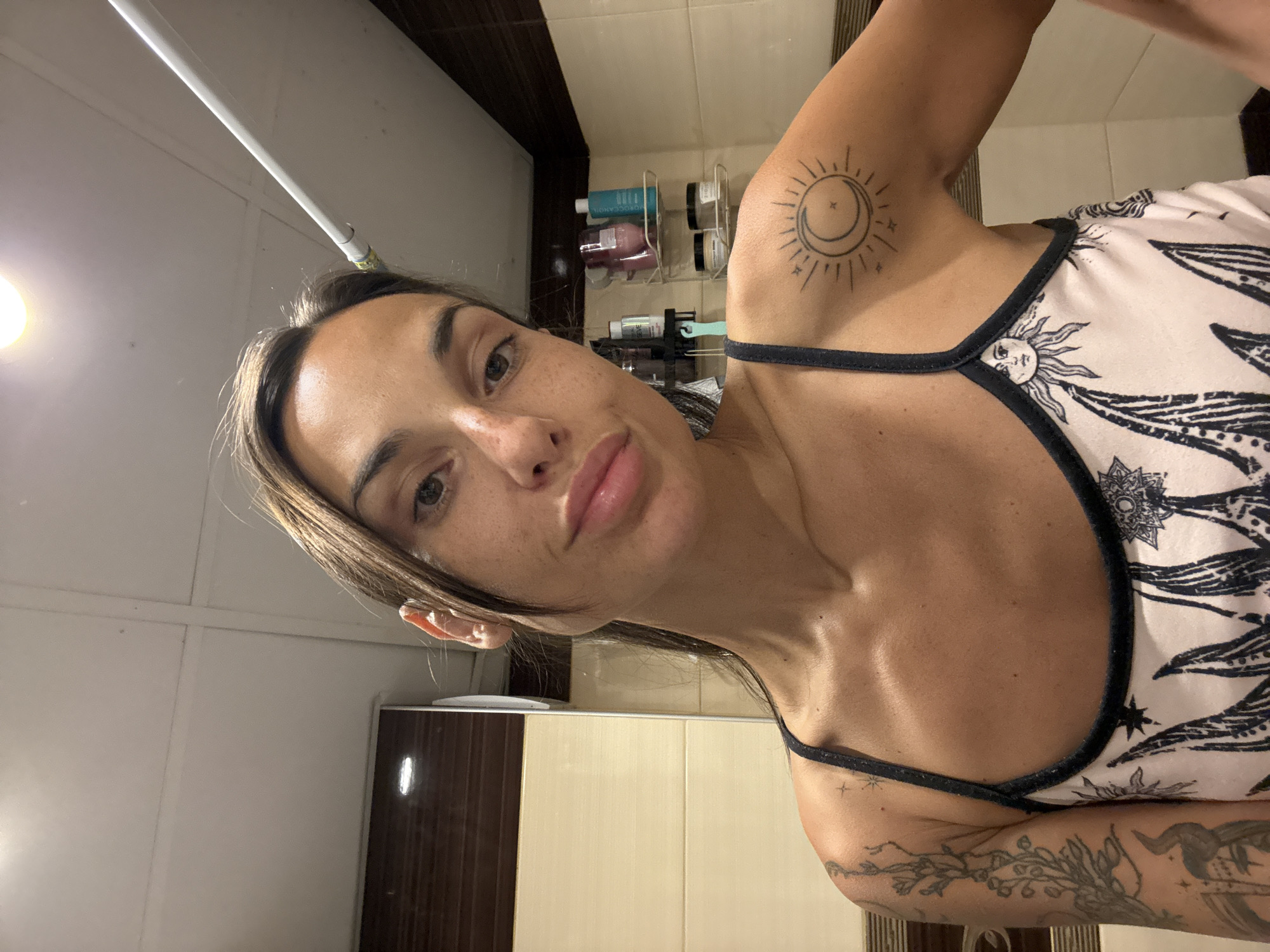 A woman with brown hair and freckles is taking a selfie in a bathroom. She is wearing a spaghetti strap top with a celestial print. She has tattoos on her left arm and right shoulder. The bathroom has tiled walls and shelves with various toiletries. There is a light fixture on the ceiling. The image does not provide any information about the location within the city of Pleven, Bulgaria.