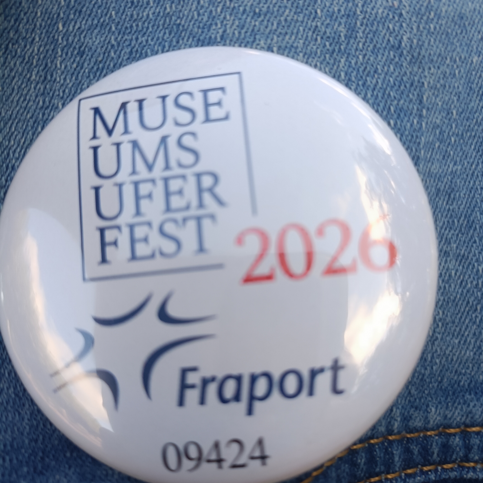 The image shows a close-up of a white circular pin badge with text and graphics on it, placed on blue denim fabric. The badge has a blue-outlined square containing the words "MUSE UMS FEST" in large blue capital letters. To the right of this text, the numbers "2026" are printed in red. Below the square and to the left, there are two abstract blue curved shapes. Below these shapes and centered on the badge are the words "Fraport" in blue capital letters. At the very bottom of the badge, the number "09424" is printed in dark gray. The background is denim, suggesting the badge is pinned to clothing. Given the presence of "Fraport," which is the operator of Frankfurt Airport, the badge likely relates to an event or organization associated with Frankfurt, Germany.