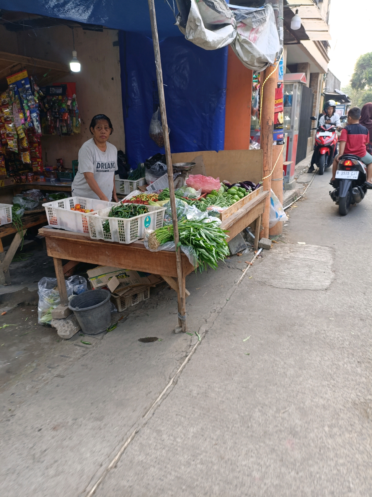 The image depicts an outdoor market stall in Tangerang, Indonesia, likely on a street or alley. A woman, appearing to be the vendor, stands behind a wooden table laden with fresh produce. She is wearing a light-colored t-shirt with a graphic design. The table displays various vegetables in white plastic baskets, including tomatoes, chilies, and green leafy vegetables. To the left of the stall, shelves are stocked with packaged goods. In the background, a busy street is visible, with a motorcycle carrying two people driving away from the camera. The overall scene suggests a vibrant local marketplace.
